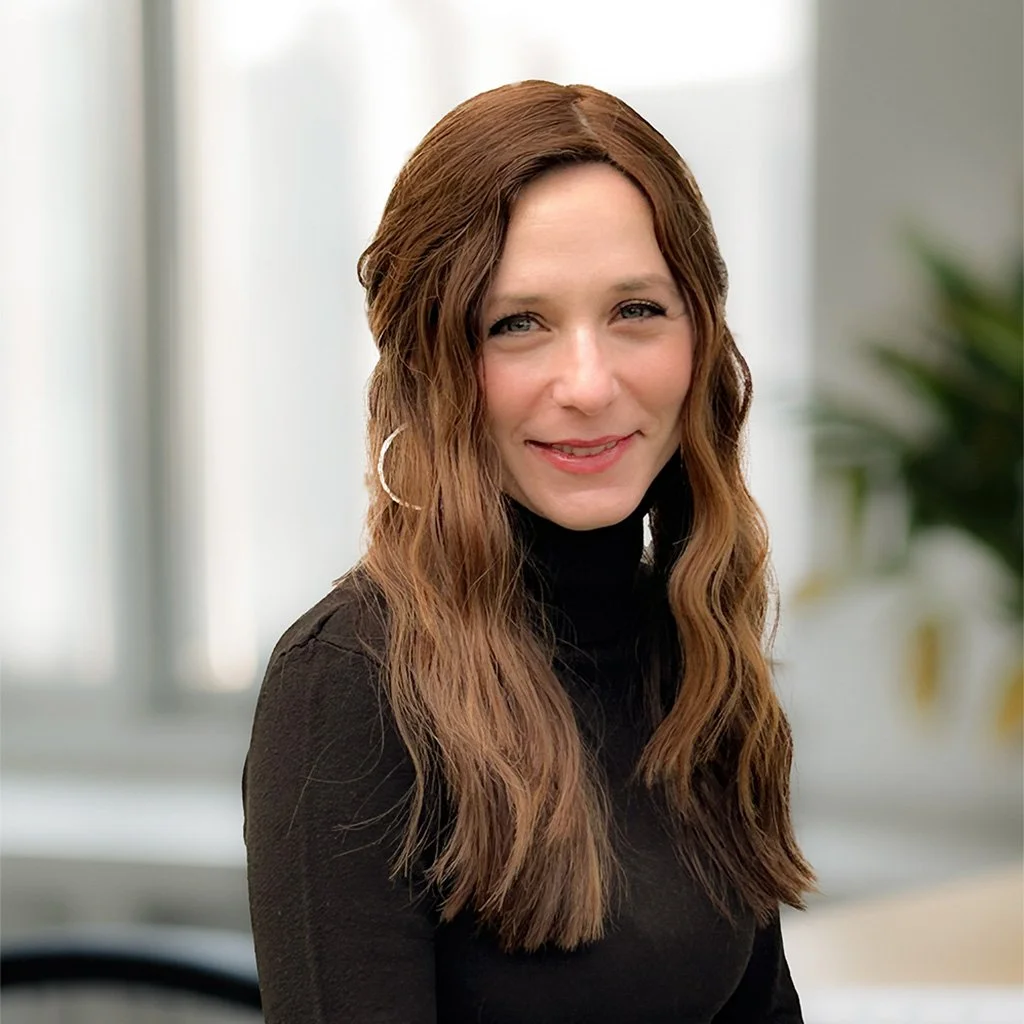 A woman with long wavy brown hair, wearing hoop earrings and a black turtleneck, smiling at the camera in an indoor setting.