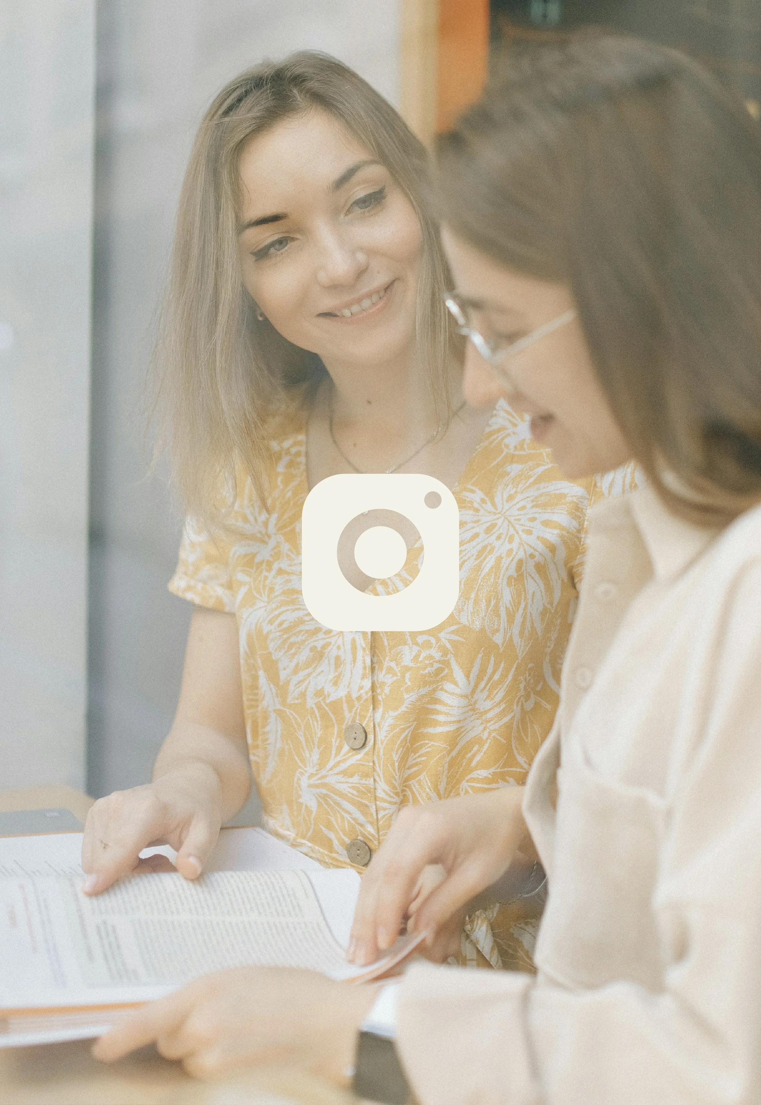 Two women are engaged in a discussion, looking at a book or document together. One woman has long, light brown hair and is wearing a yellow and white patterned shirt. The other woman has dark brown hair, glasses, and is wearing a light-colored blouse.