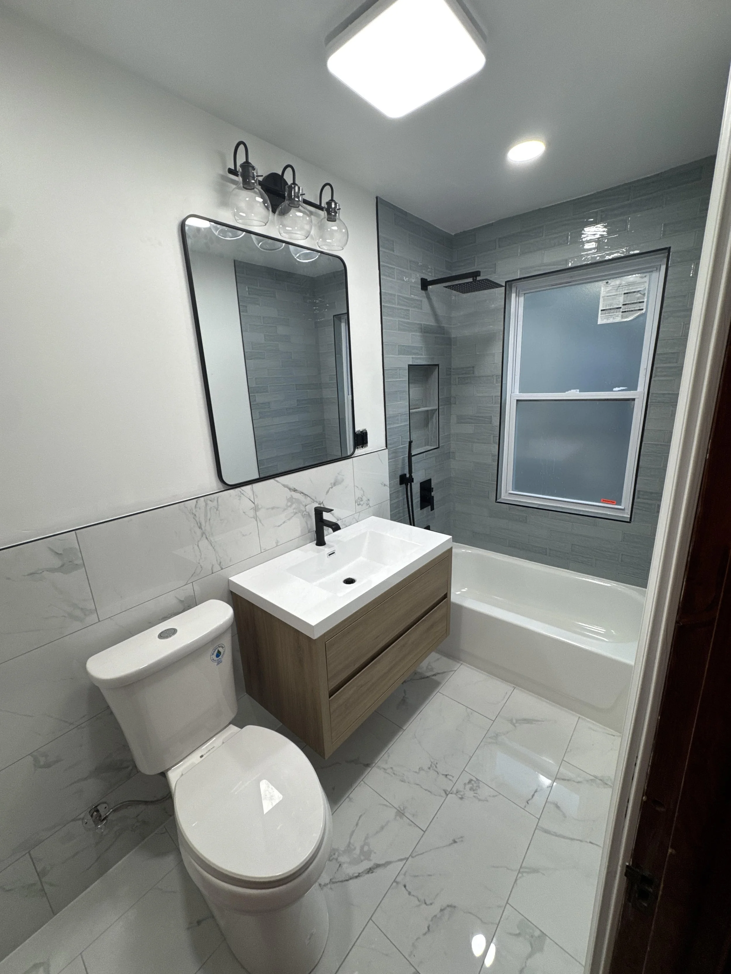 Modern bathroom with white marble floor, white toilet, wooden vanity with white sink, large mirror, black fixtures, gray tile shower with glass door, window, and a ceiling light.