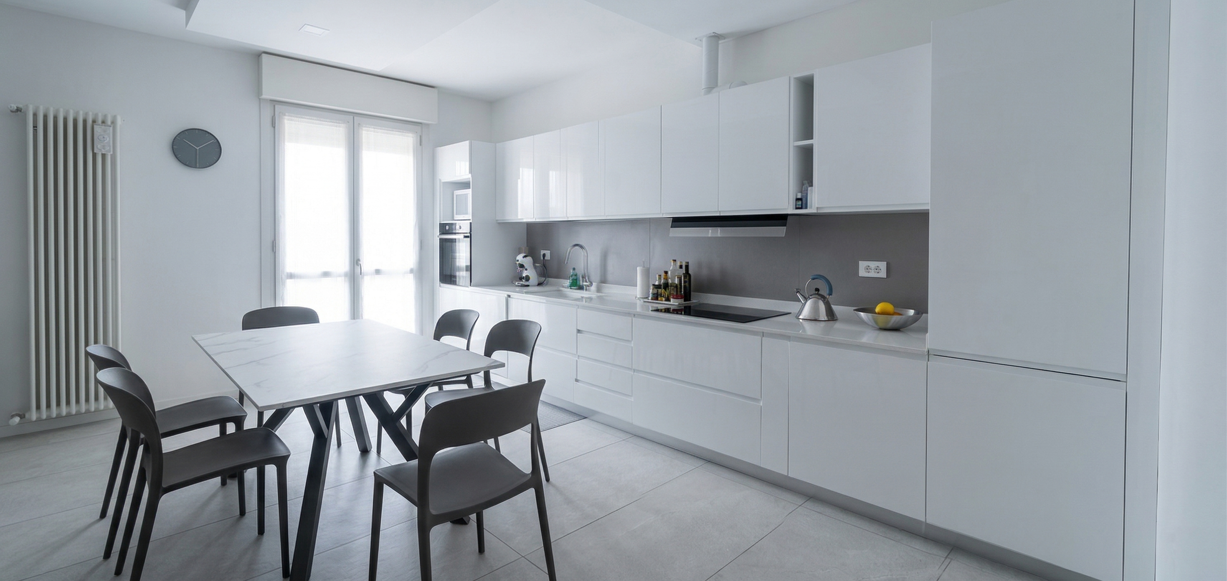 Cucina moderna bianca con tavolo e sedie grigie, grandi finestre con tende bianche, orologio a parete, radiatore bianco, e vari elettrodomestici e oggetti da cucina.