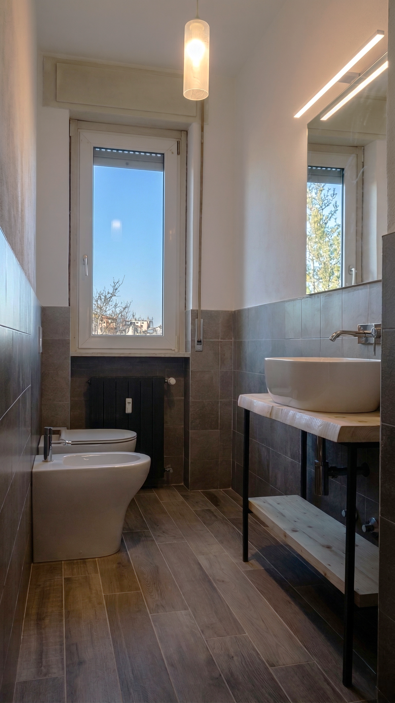 Bagno moderno con pareti piastrellate, water e bidet, finestra con vista su cielo e alberi, lavabo su mensola di legno con supporto in metallo, illuminazione a incasso e a sospensione.