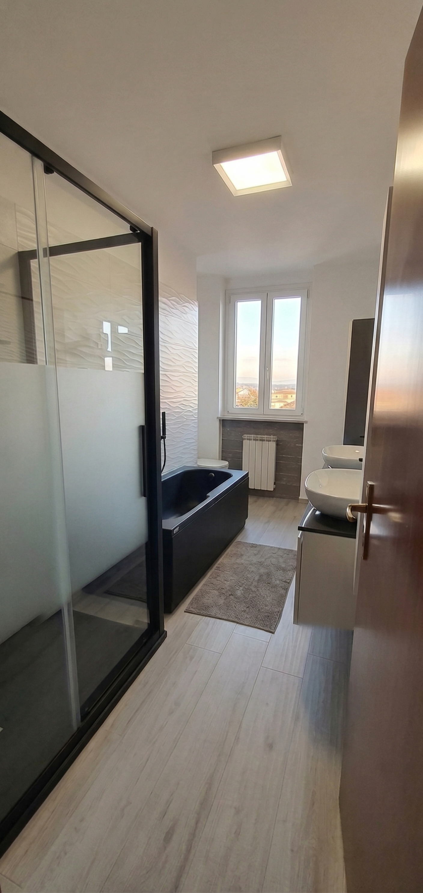 Bagno moderno con vasca nera, doccia con porte in vetro, doppio lavabo a bassorilievo, finestra e termosina. Pavimento in piastrelle chiare.