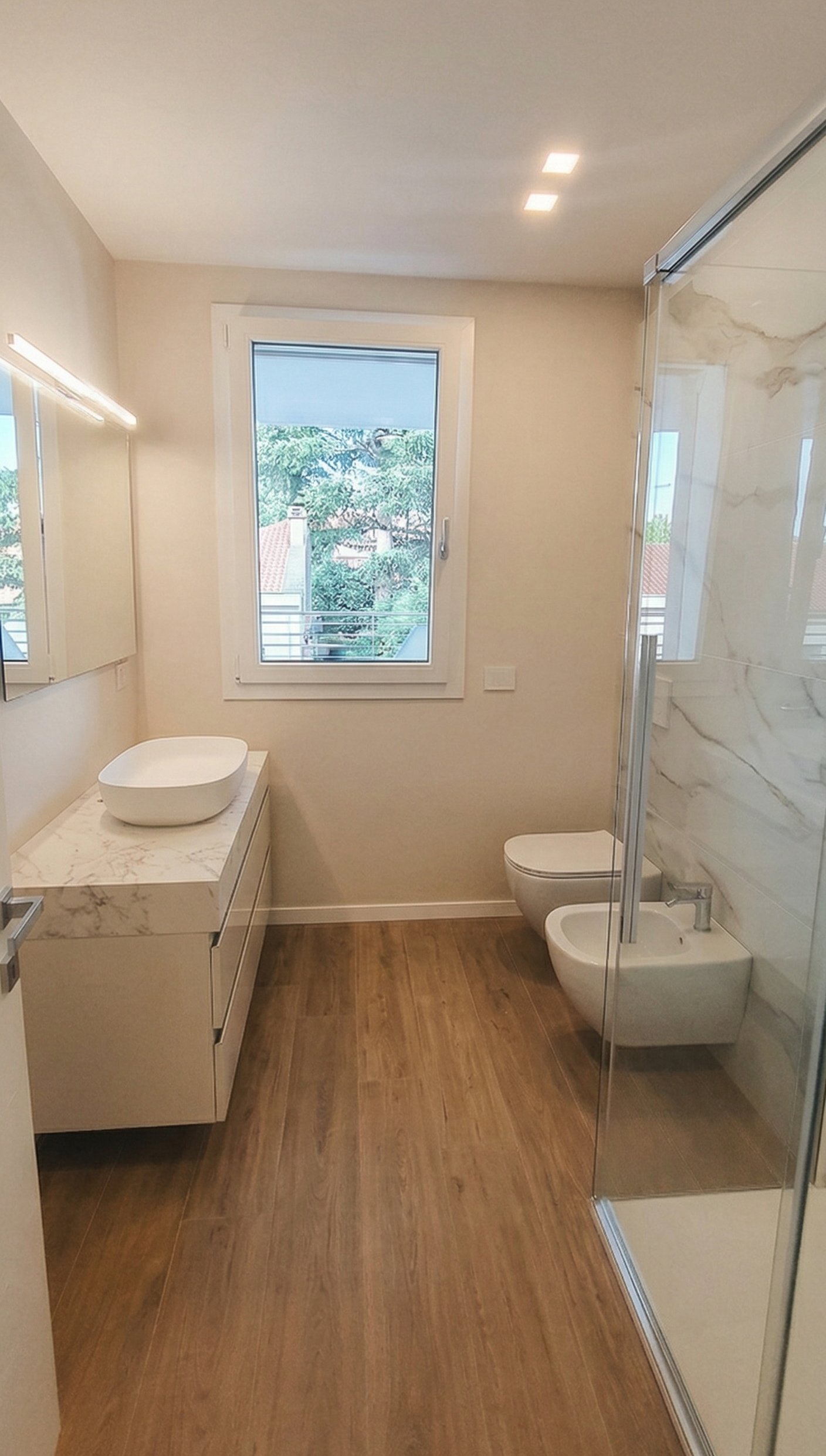 Bagno moderno con mobili bianchi, lavabo su terrazzo con venature marmo, WC, bidet, doccia con pareti in vetro, finestra con vista su alberi e case, illuminazione a soffitto.