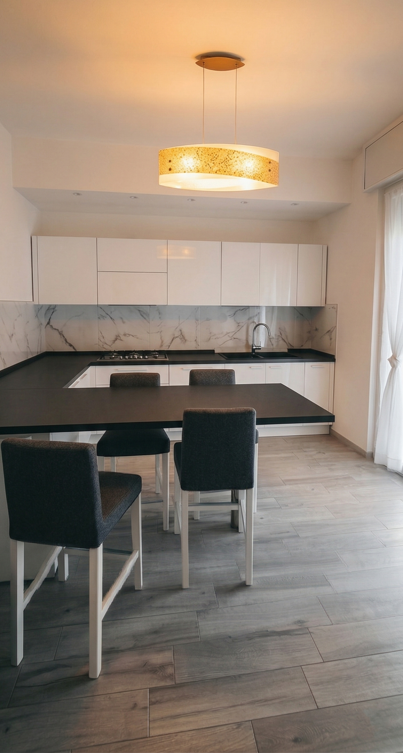 Cucina moderna con piano di lavoro nero, mobili bianchi, parete in marmo e un tavolo con sedie nere. Lampada a soffitto rotonda gialla. Luminosità naturale da una finestra con tende bianche.