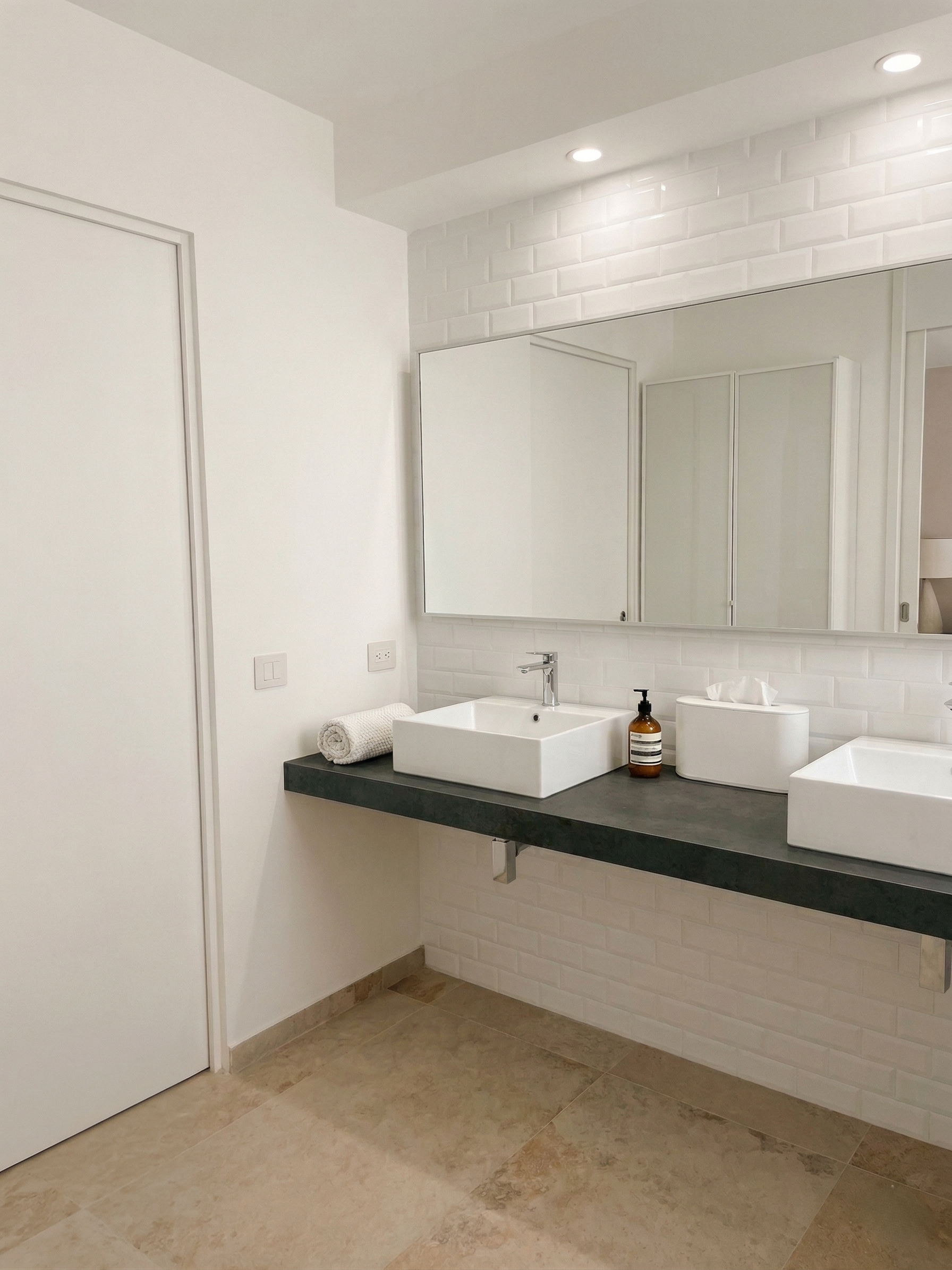 Bagno con lavabo bianco a forma di scatola, specchio grande, detergente in bottiglia marrone, scatola di fazzoletti, asciugamano bianco arrotolato, pareti bianche, piastrelle beige, luci incassate nel soffitto.