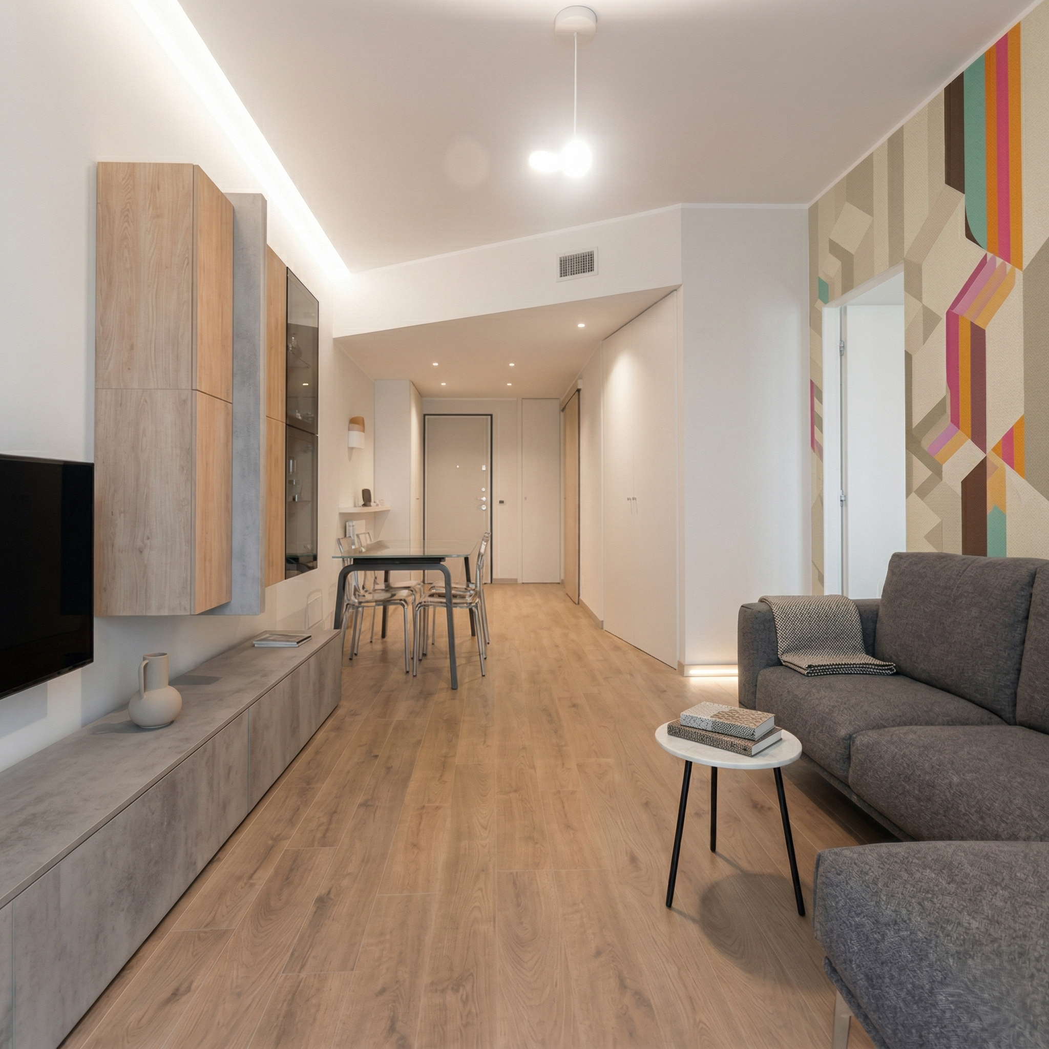 Interno di un soggiorno moderno con parete attrezzata in legno, divano grigio, tavolino con libri, parete decorativa colorata, e tavolo da pranzo con sedie. Pavimento in legno chiaro.