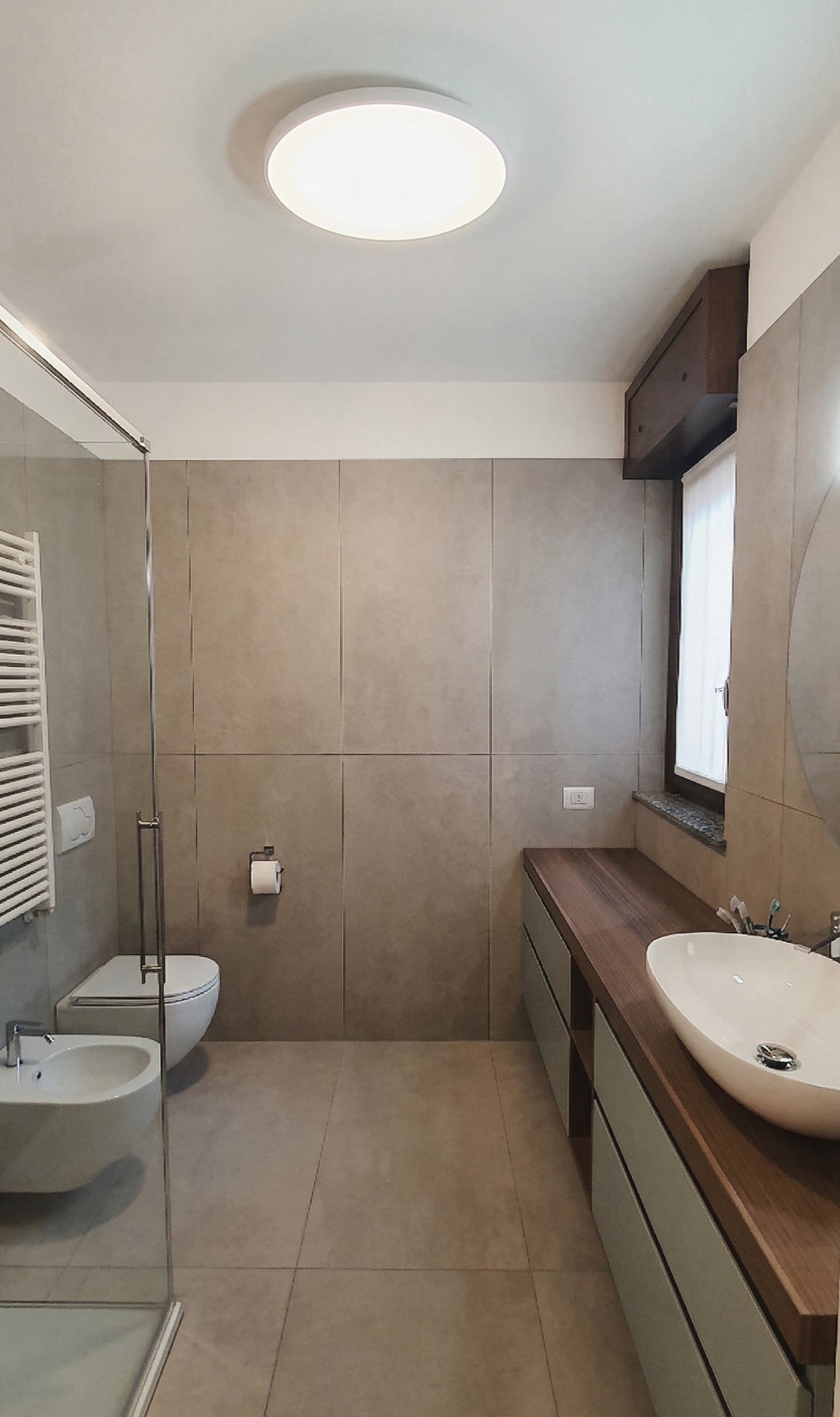 Bagno moderno con pareti e pavimenti rivestiti di piastrelle beige, grande specchio con dettaglio curvo, lavabo ovale bianco su mobile in legno scuro, finestra con tende bianche, doccia in vetro, tenda riscaldante bianca, inserto elettrico sulla pare