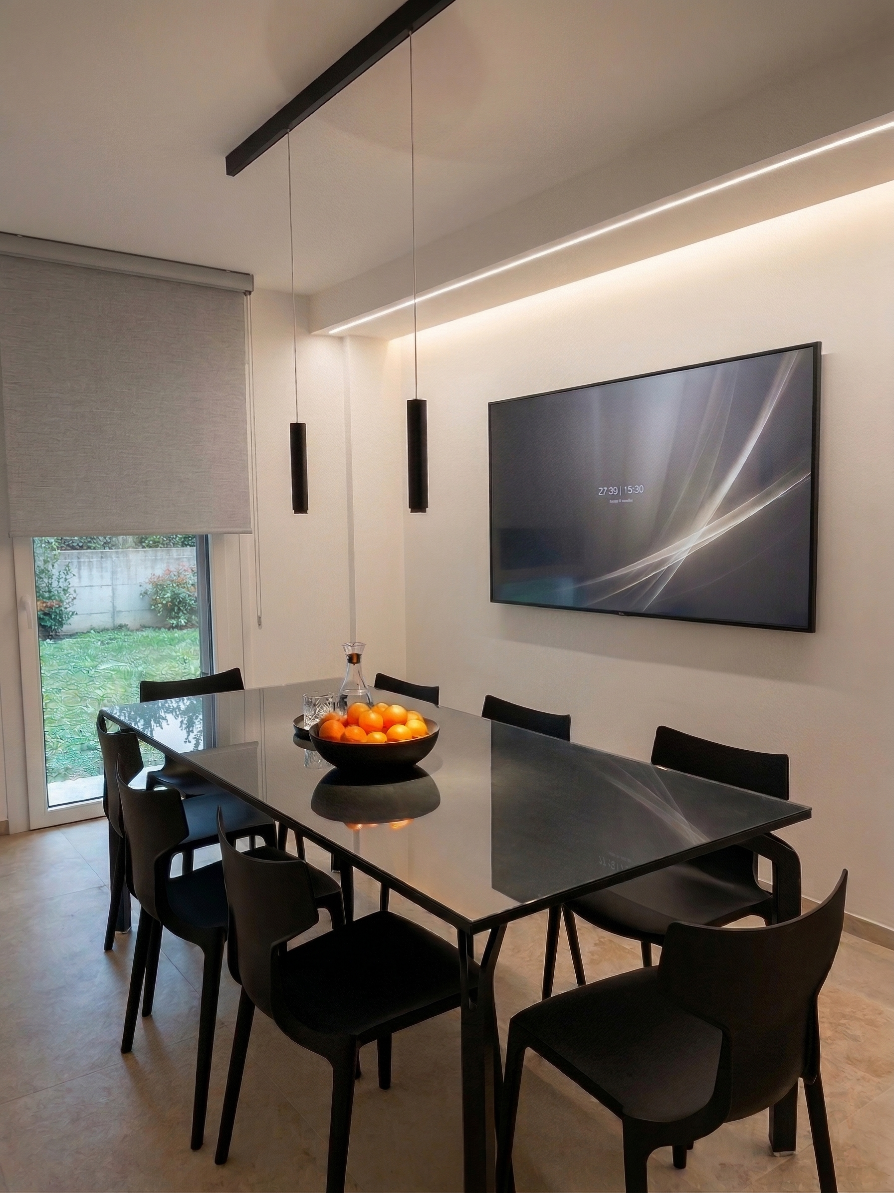 Sala da pranzo moderna con tavolo in vetro nero, sedie nere, un grande schermo TV sulla parete, un vassoio di arance e una brocca d'acqua centrale, lampade sospese nere e una finestra con tenda