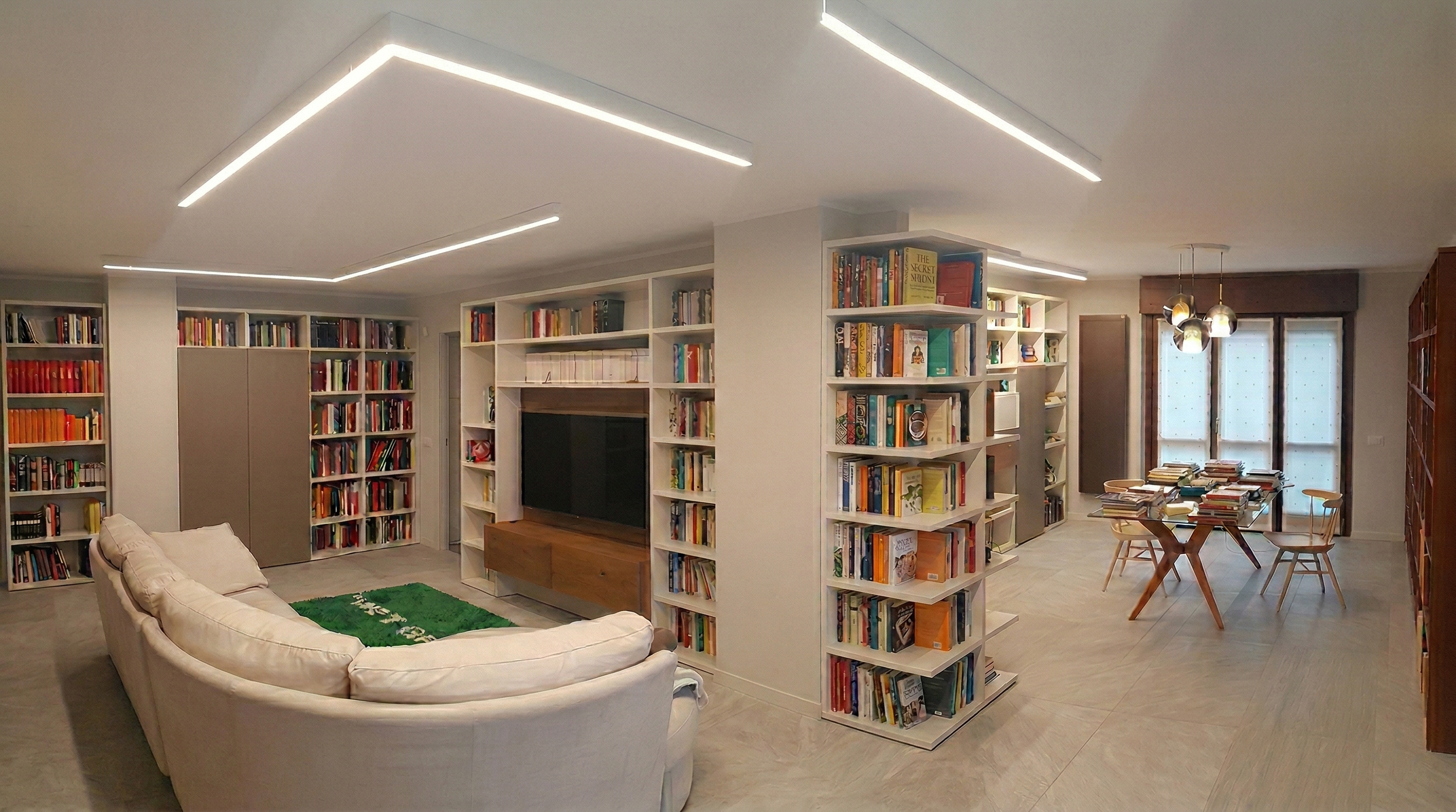 Ampia zona soggiorno con divano bianco, librerie cariche di libri, area lettura con tavolo e sedie, e un televisore