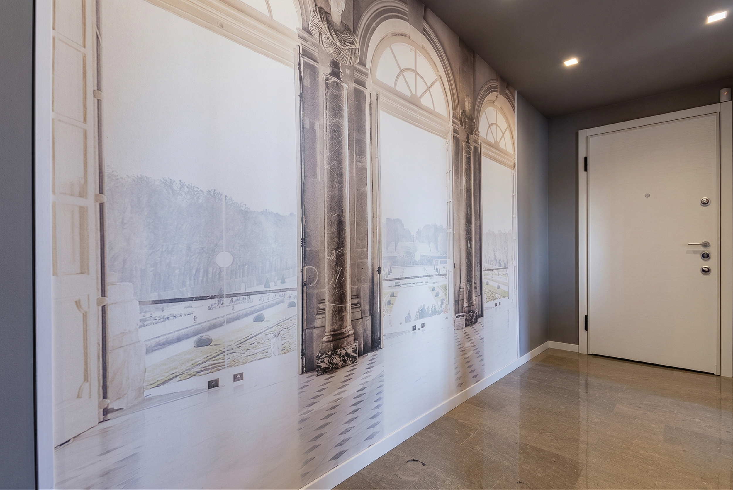 Muro decorato con un'illustrazione di una grande finestra con archi e colonne in stile classico, che mostra una scena di un giardino con alberi e un lago, nella parte superiore dell'ingresso di una casa.