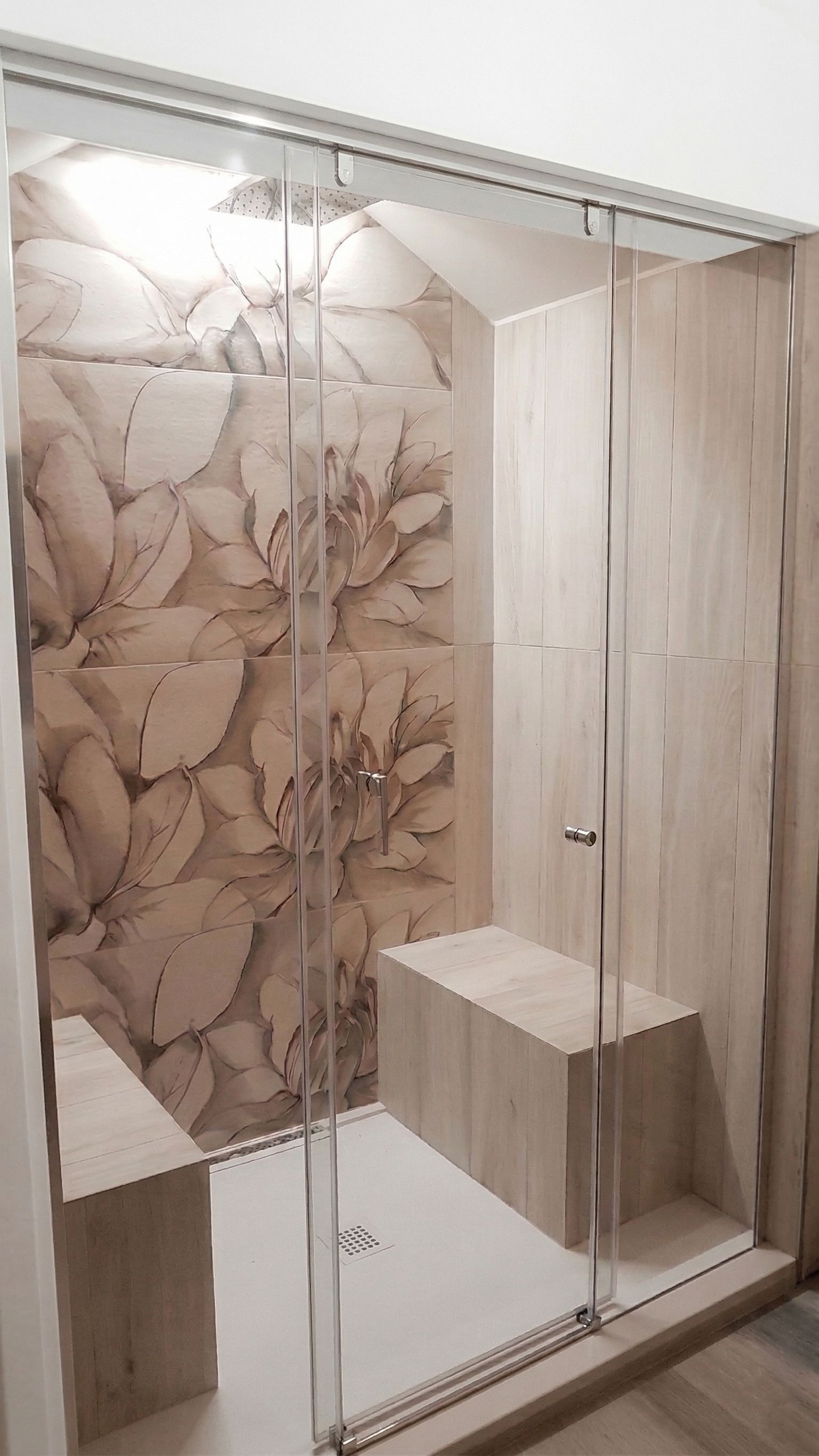 Doccia moderna con pareti in vetro, rivestimenti in piastrelle beige con motivi floreali e inserti in legno, con due sedili integrati.