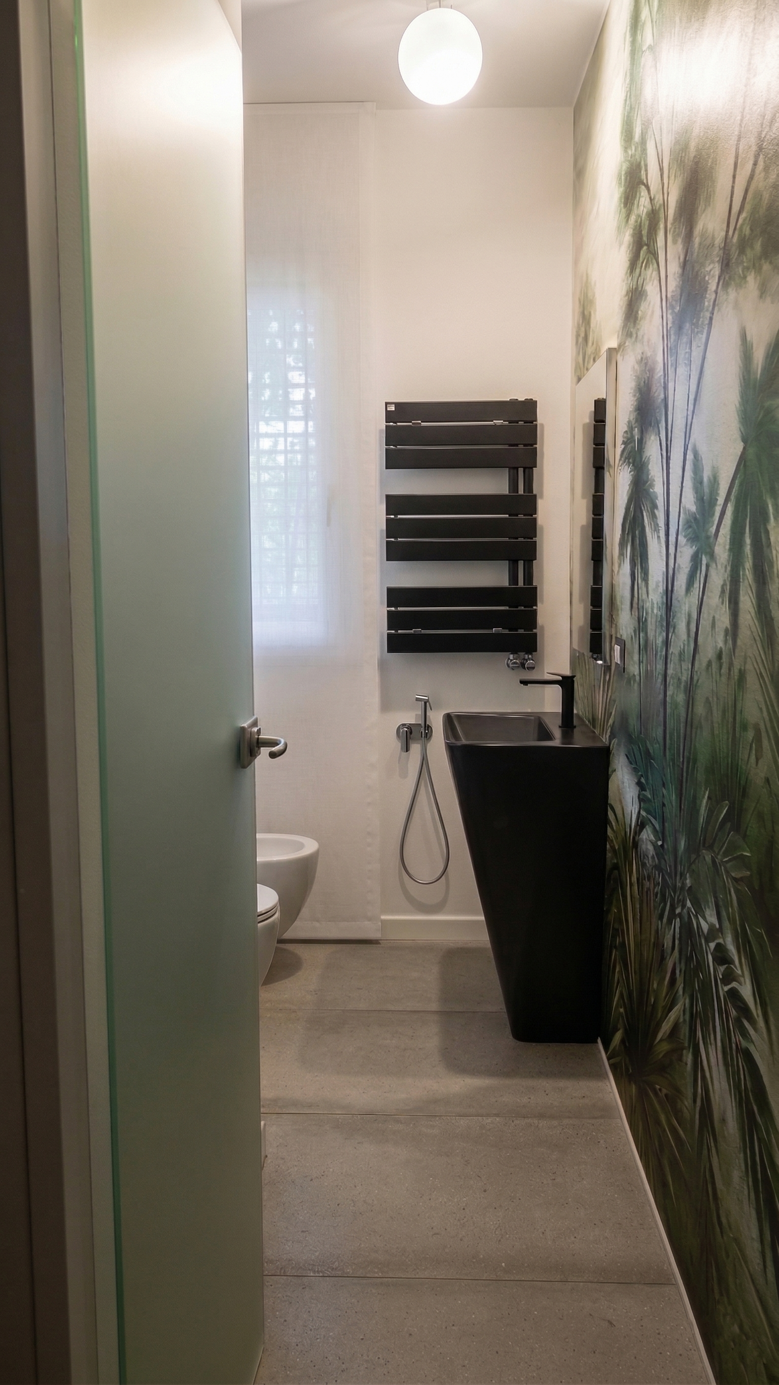 Una stanza da bagno moderna con pareti decorate con una carta da parati a stampa foglia di palme, un lavabo nero e uno specchio, un termosifone nero e un water visibile tra una porta aperta e una finestra con tende trasparenti.