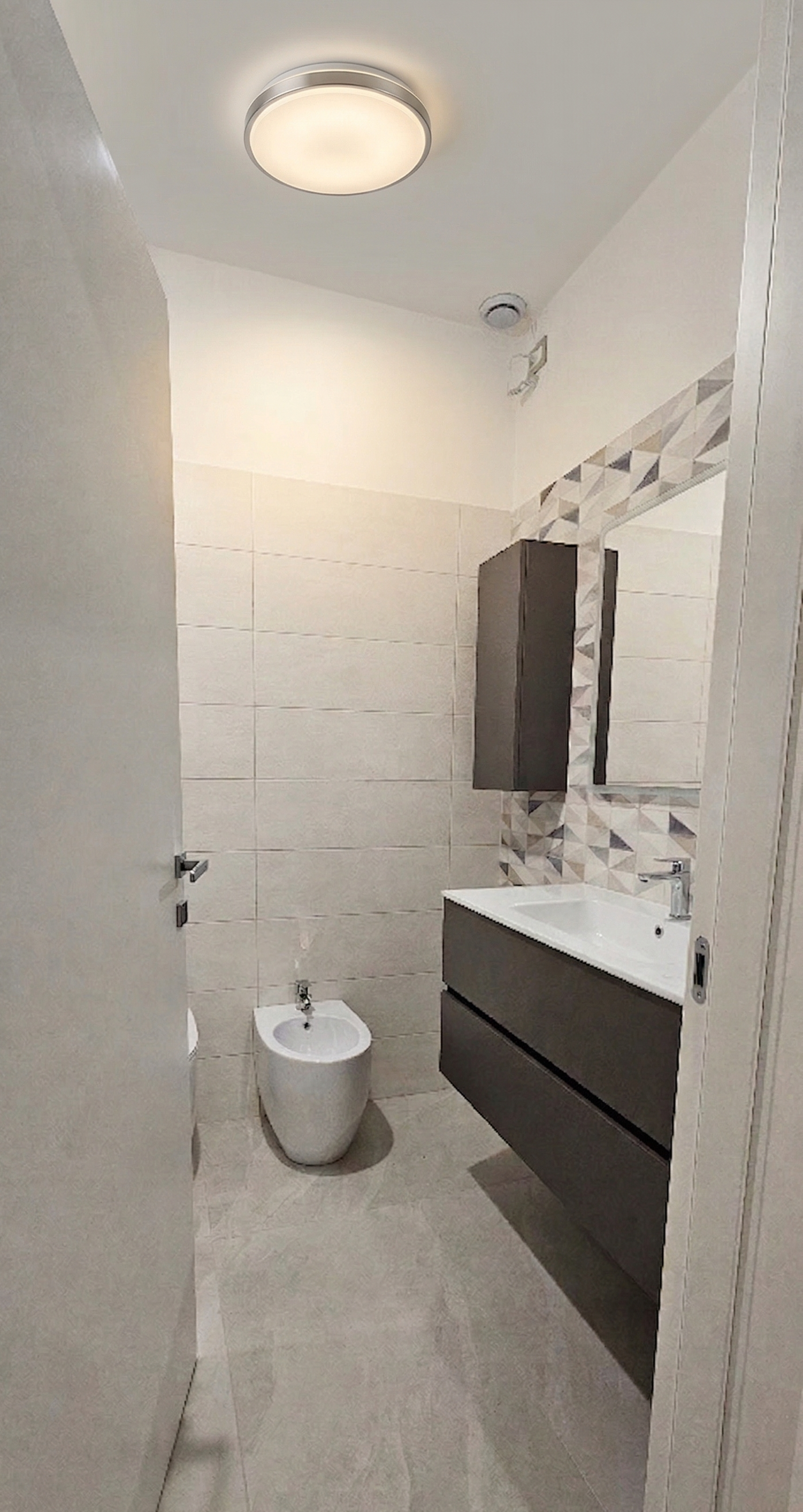 Bagno con pareti rivestite con piastrelle beige, lavabo con mobile portaoggetti grigio scuro, specchio e armadietto sopra il lavabo, luci a soffitto, bidet e parete con motivo decorativo di piastrelle geometriche.