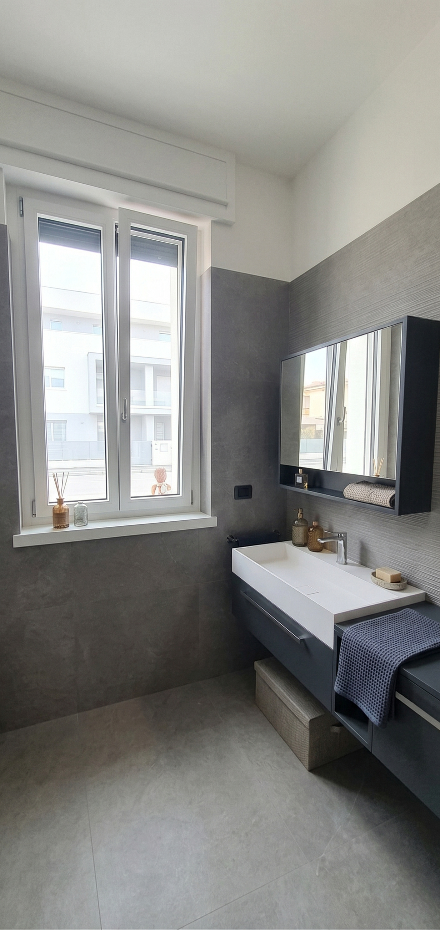 Bagno moderno con finestra, lavabo bianco con arredi neri, specchio grande, zona con piccoli contenitori per sapone e accessori, tenda grigia a pois e oggetti decorativi sulla finestra.