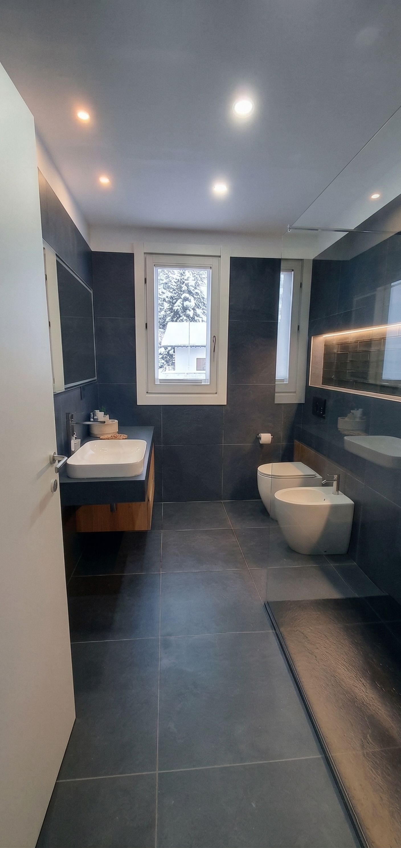 Bagno moderno con pareti e pavimenti in piastrelle grigie scure, lavabo con mobile in legno, wc e bidet bianchi, finestra con vista sulla neve e alberi, specchio con illuminazione integrata, portaspugnetta e porta
