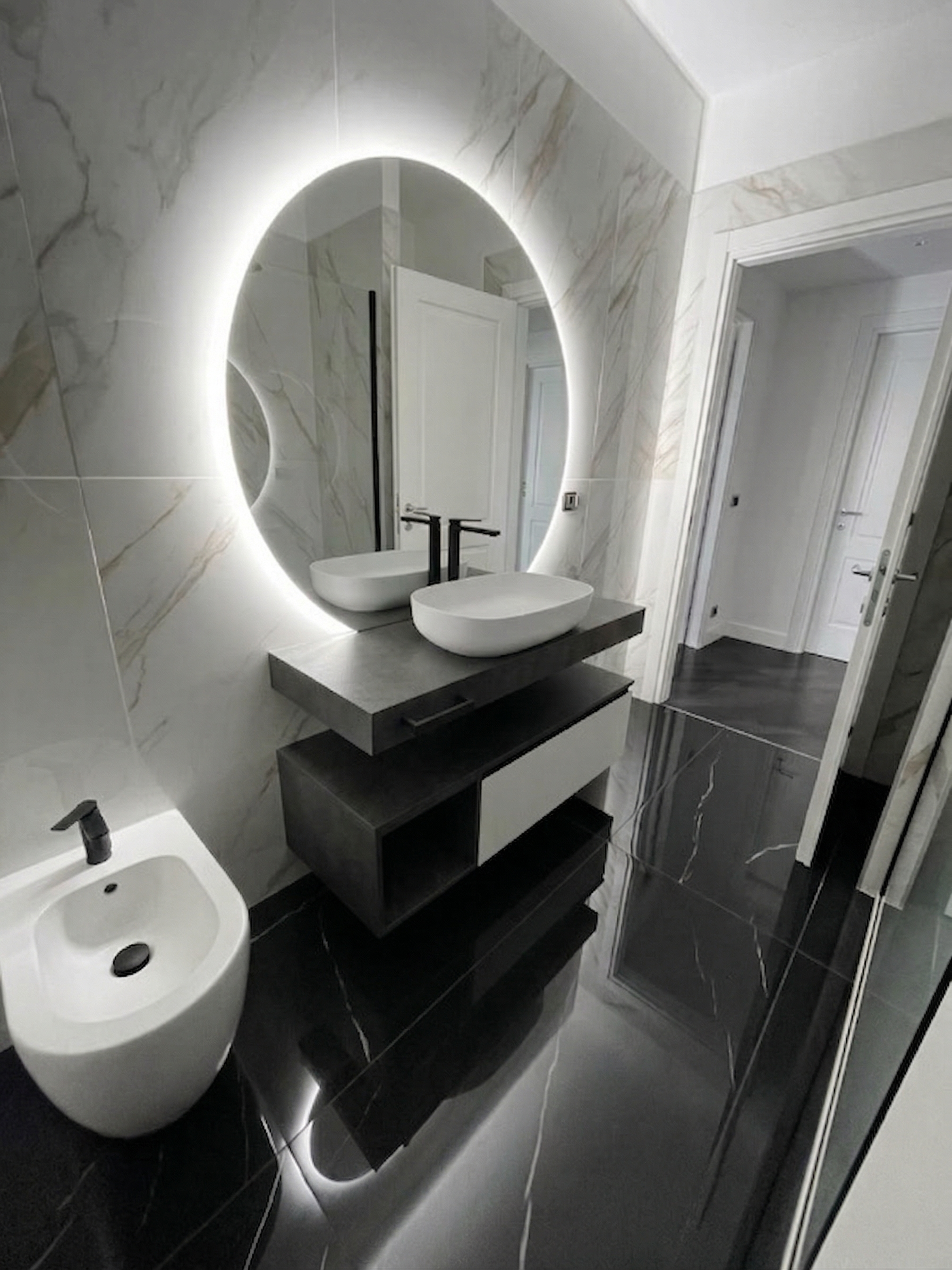 Bagno moderno con pareti in marmo chiaro, lavandino rotondo sopra un mobile nero e bianco con uno specchio rotondo con illuminazione a LED.