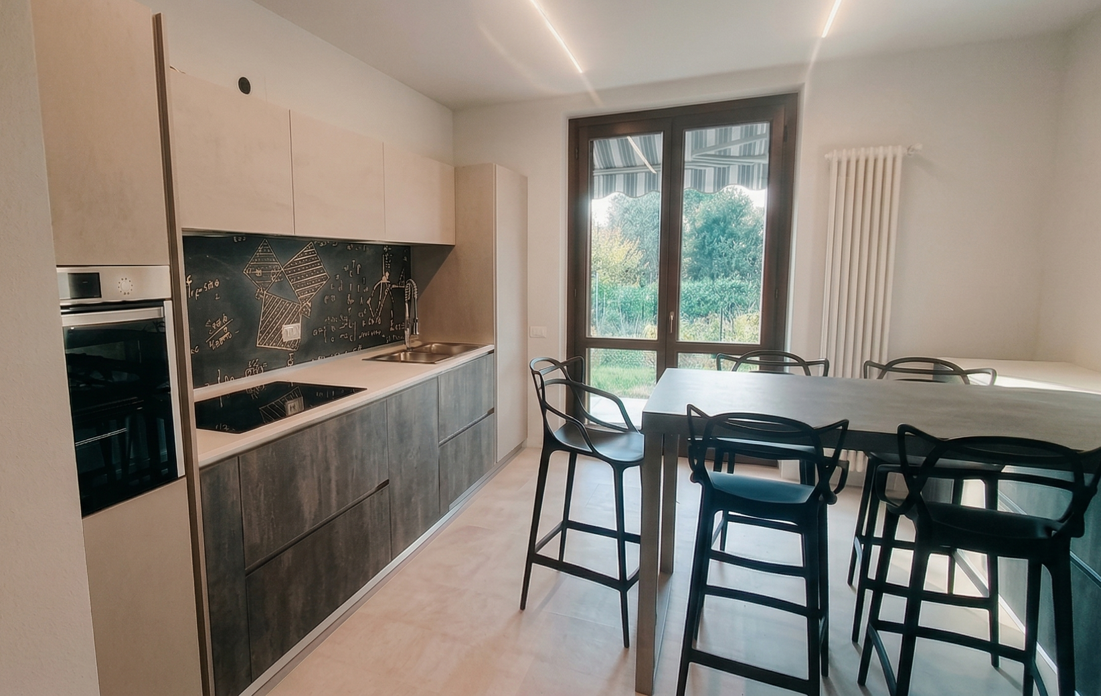 Cucina moderna con ante in legno chiaro e parte inferiore in finitura grigia, piano di lavoro bianco, parete con lavagna nera decorata con disegni e scritte, grande finestra con vista su un giardino verde, tavolo bianco con sedie nere.