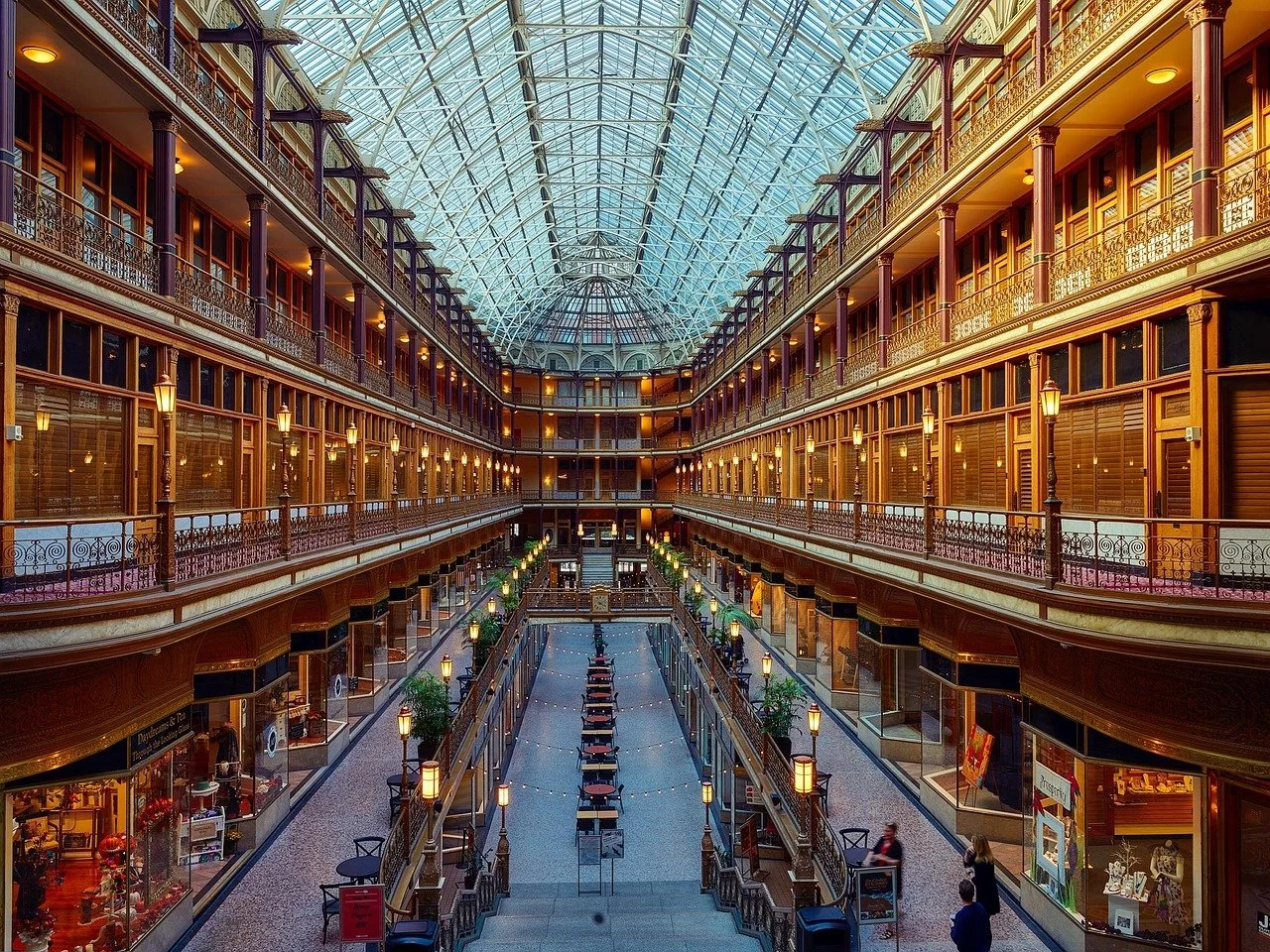 Interior of a large shopping mall with multiple floors, ornate wooden railings, and a glass-domed ceiling. The ground level has storefronts and a central corridor with seating and decorative street lamps.