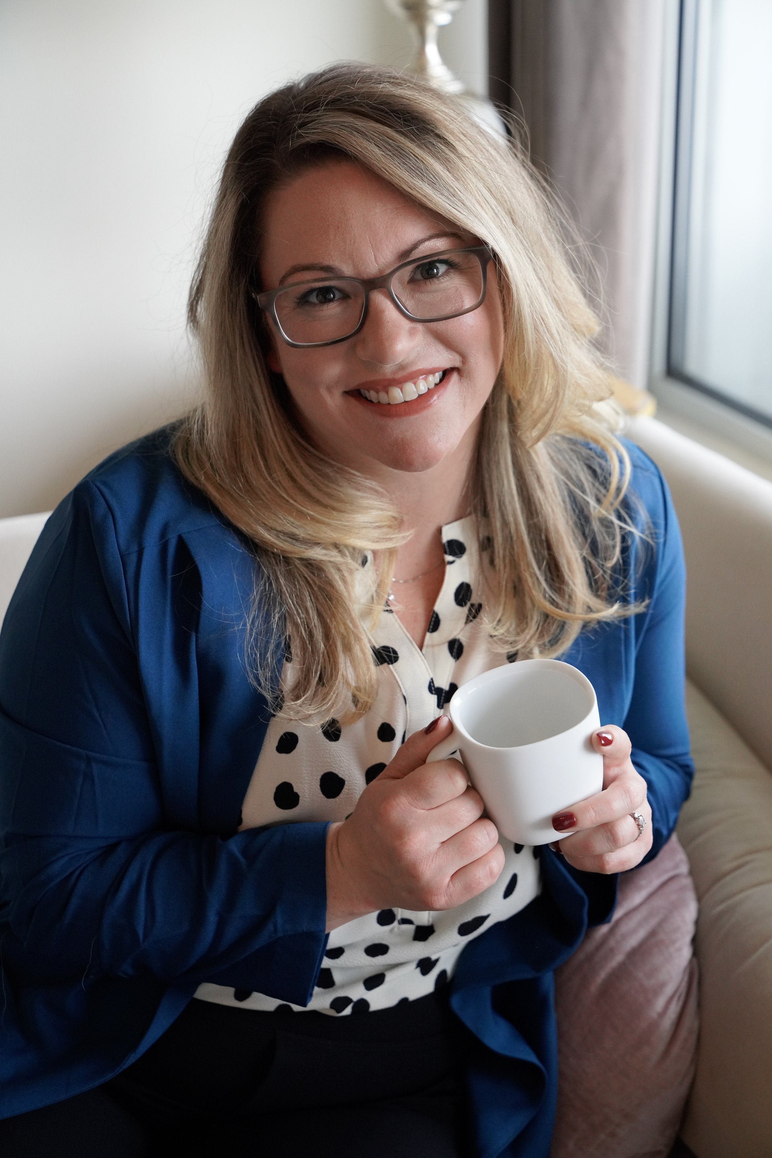 A woman with glasses and blonde hair, smiling, holding a white mug, sitting on a beige sofa next to a window.