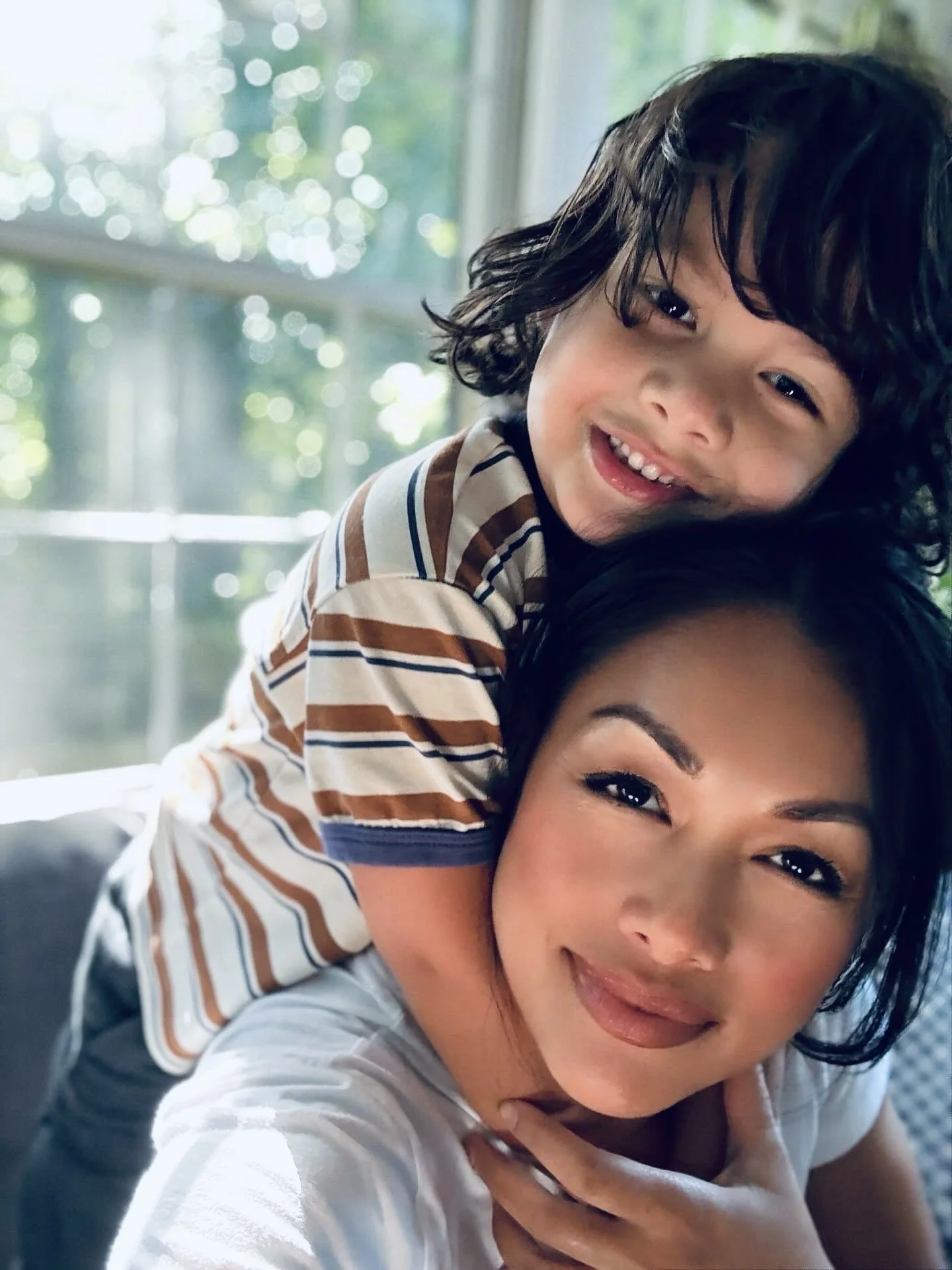 A woman and a young boy smiling, with the boy on her back inside a room with a window and greenery outside.
