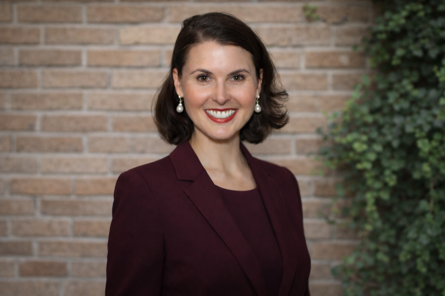 A woman with shoulder-length dark hair smiling, wearing a burgundy blazer and pearl earrings, standing in front of a brick wall with some greenery on the side.