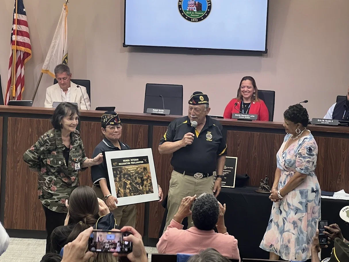 A recognition ceremony honoring women veterans, featuring five women standing in front of a panel with people seated behind it. One woman holds a framed poster titled 'Women Veteran Recognition Proclamation.' Several people in military hats are prese