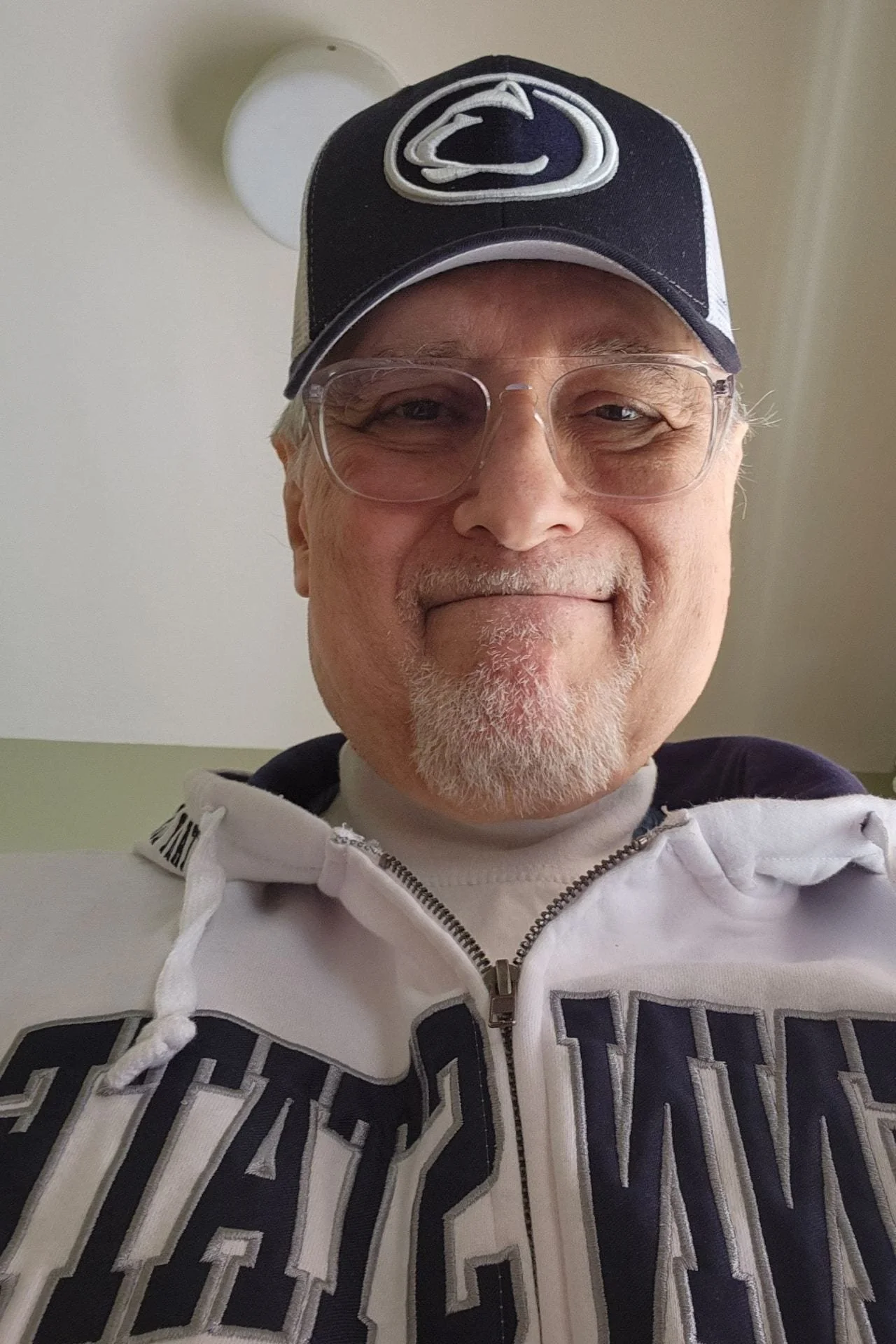 Close-up of an older man wearing glasses, a Philly Phillies baseball cap, and a zip-up jacket with the team's logo visible.