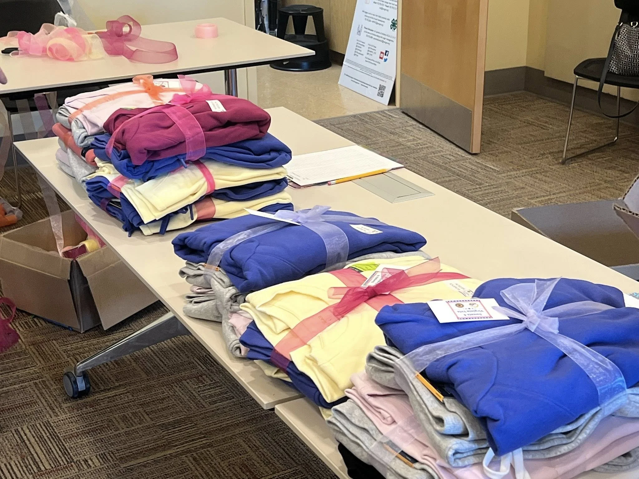 Stacked colorful clothing on a table, with ribbons and tags, at an indoor event or donation center.