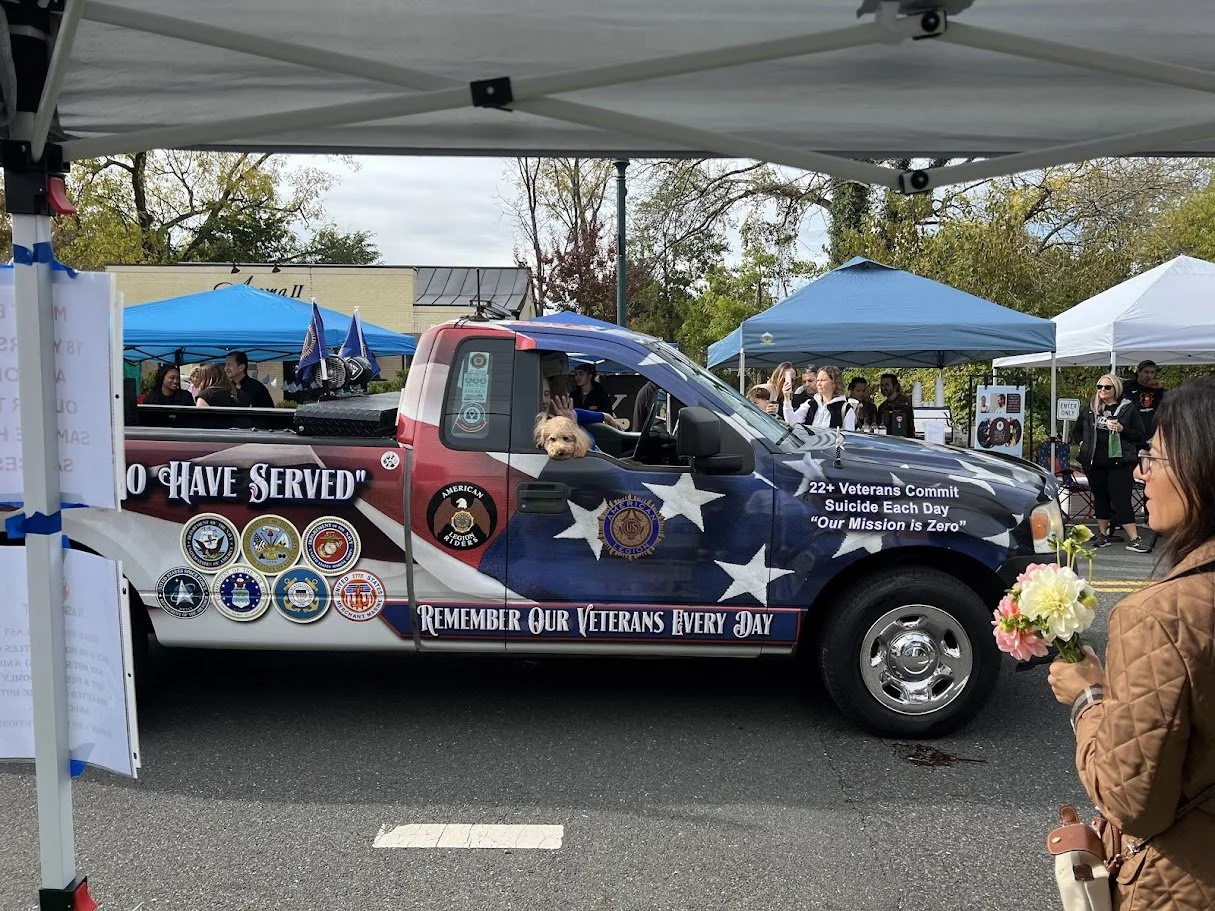 A decorated truck with patriotic themes and Vietnam War veterans' medals parked at a memorial event. People are gathered around tents, and a woman holds a bouquet of flowers.