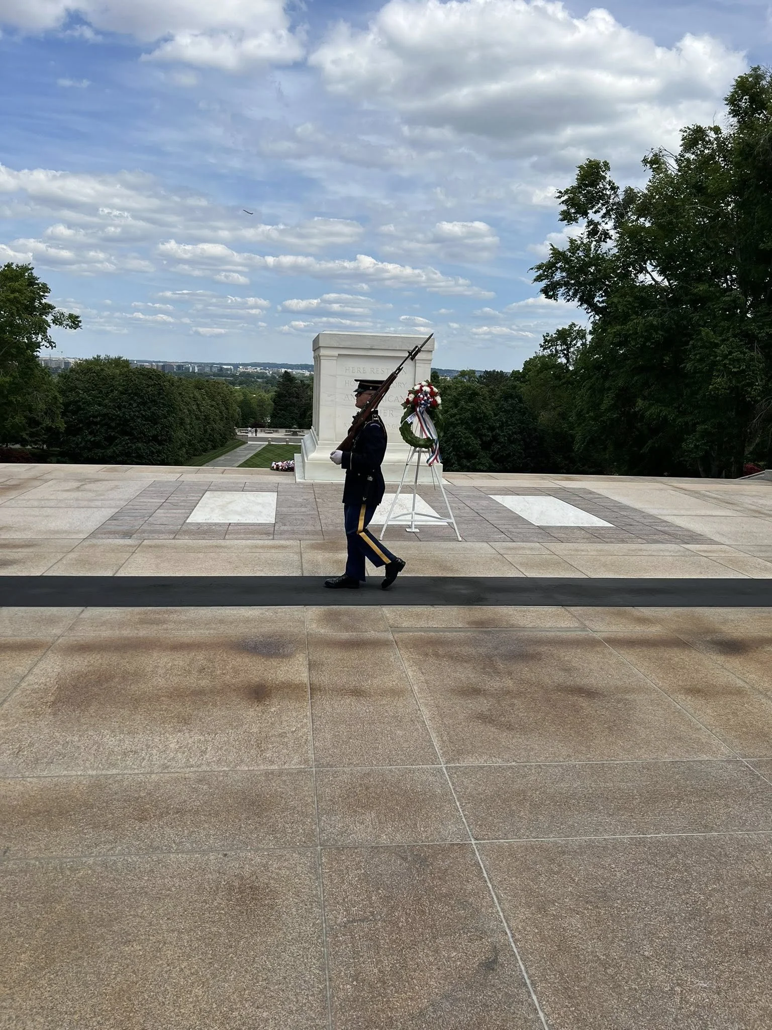 A soldier in uniform holding a rifle standing next to a wreath at a memorial site with a white monument and trees in the background.