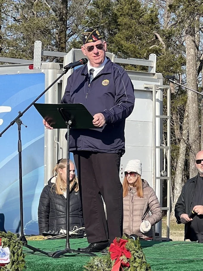 A man in a veteran's hat and sunglasses stands on a stage, speaking at a podium during an outdoor event. Three people are standing behind him with trees in the background.