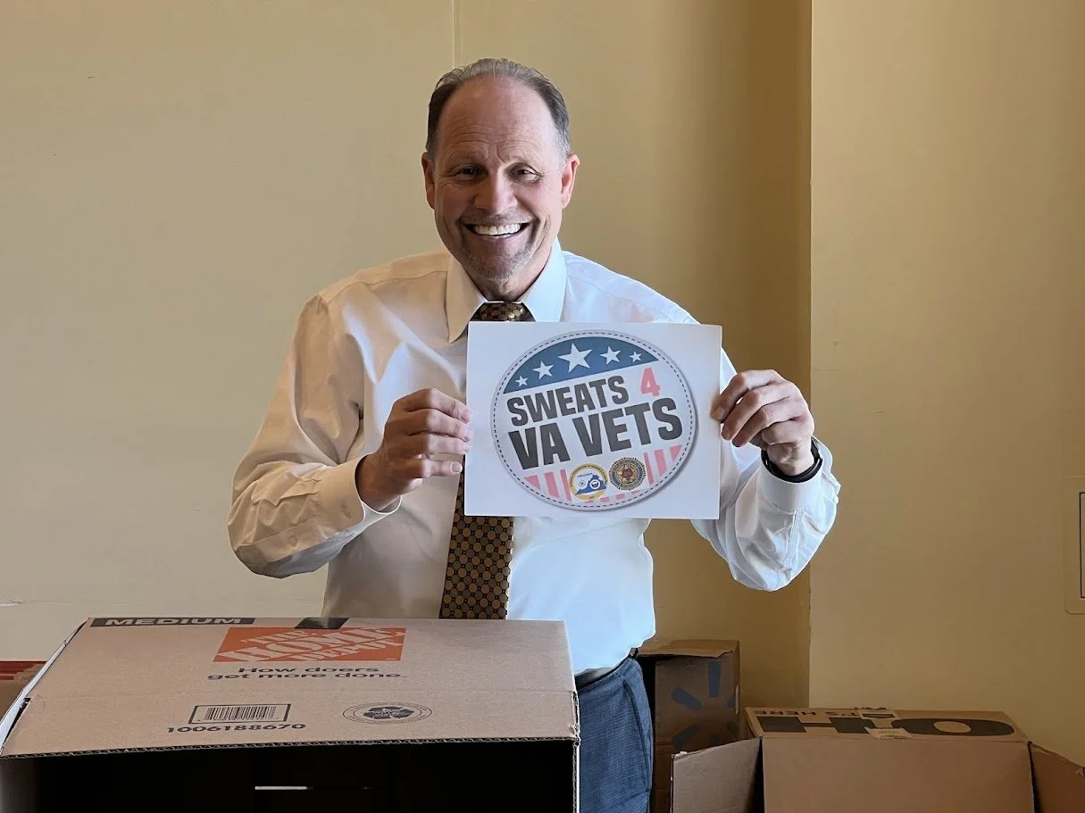 A smiling man in a white shirt and tie holding a sign that reads "Sweats 4 VA Vets" inside a room with beige walls, on a table with a cardboard box.