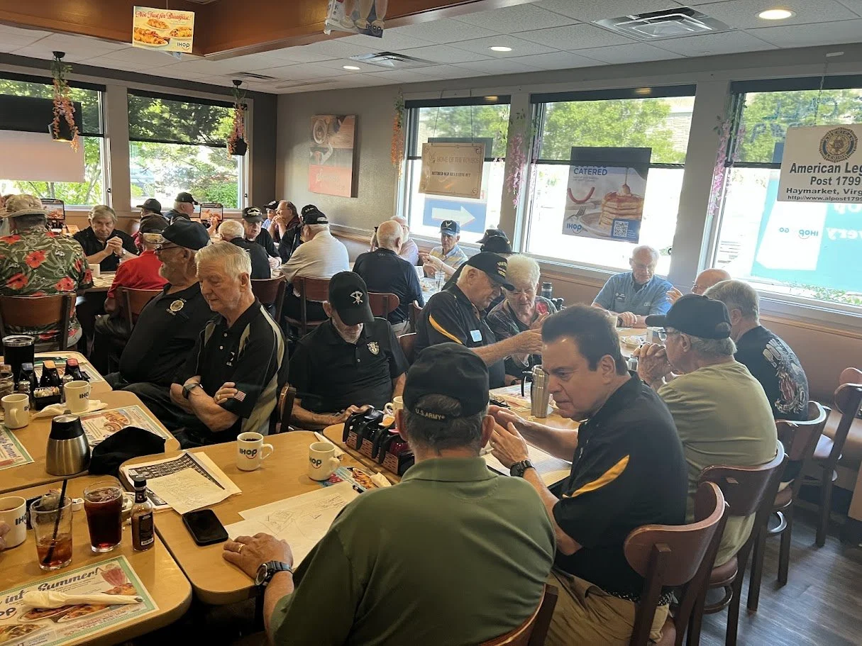 A busy IHOP restaurant filled with mostly older men, many wearing military or navy hats, seated at tables with drinks, menus, and papers, inside a well-lit room with large windows.