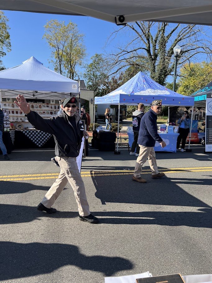 A man waving and smiling at an outdoor market with tents, people, and trees in the background.