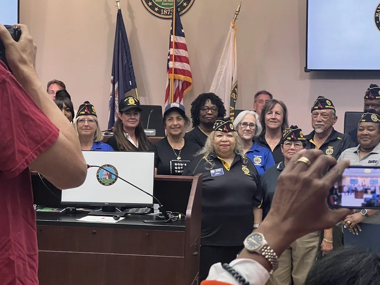 Group of veterans in uniform posing for a photo in a government meeting room with flags and a seal visible.