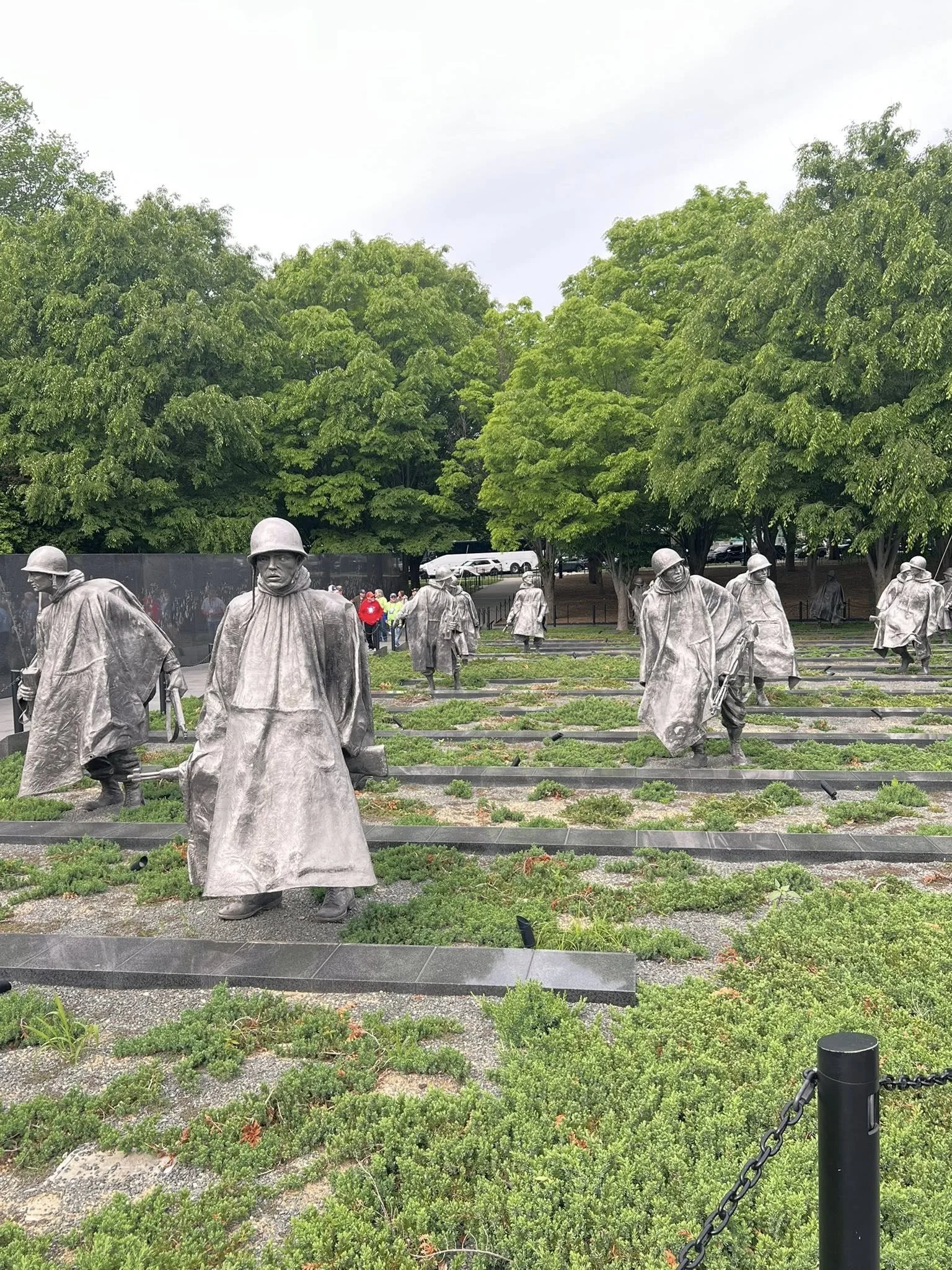 Memorial with numerous statues of soldiers in military uniforms and helmets, in a park with green trees and a black chain fence in the foreground.