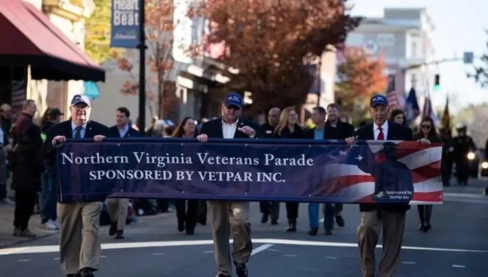 Men in suits and ties holding a banner that reads "Northern Virginia Veterans Parade SPONSORED BY VETPAR INC." during a parade on a city street with spectators and autumn trees in the background.