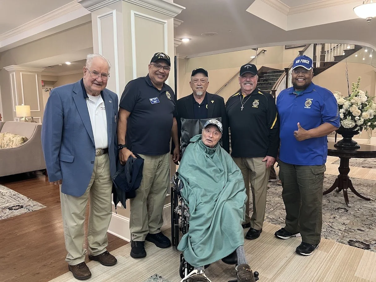 A group of six men standing around an elderly man in a wheelchair, who is covered with a green blanket. The elderly man is wearing a cap and has an surprised expression. The men are dressed casually, some in navy and black shirts, one in a blue blaze