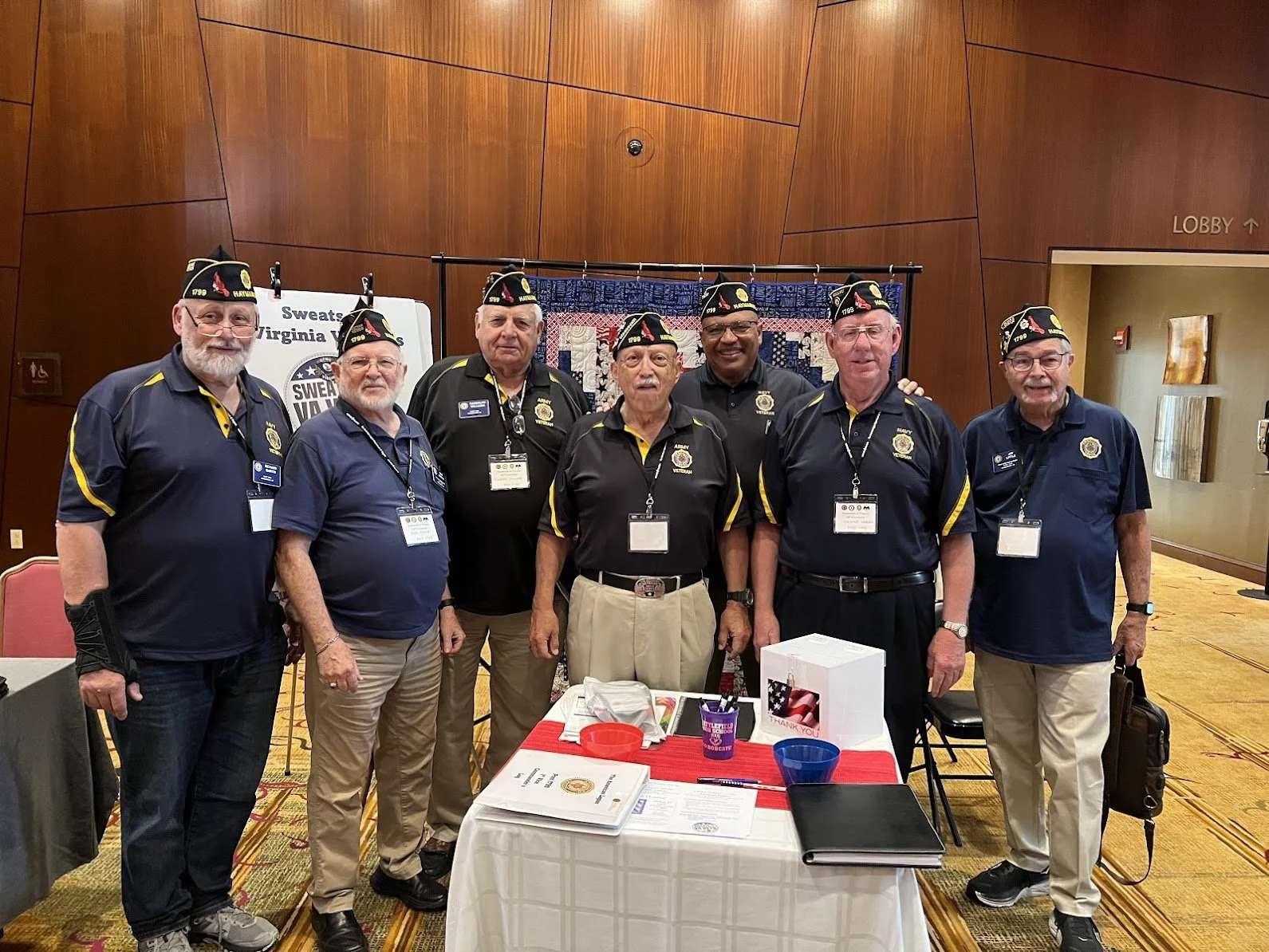 Seven men wearing veterans' organization shirts and hats, standing behind a table at an indoor event.