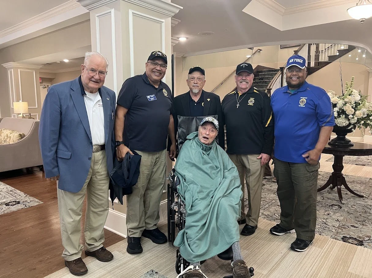 Six men stand together indoors behind an elderly man in a wheelchair, who is covered with a green blanket and wearing a cap. The men are smiling, some in police uniforms and others in casual clothing, in what appears to be a formal or celebratory gat