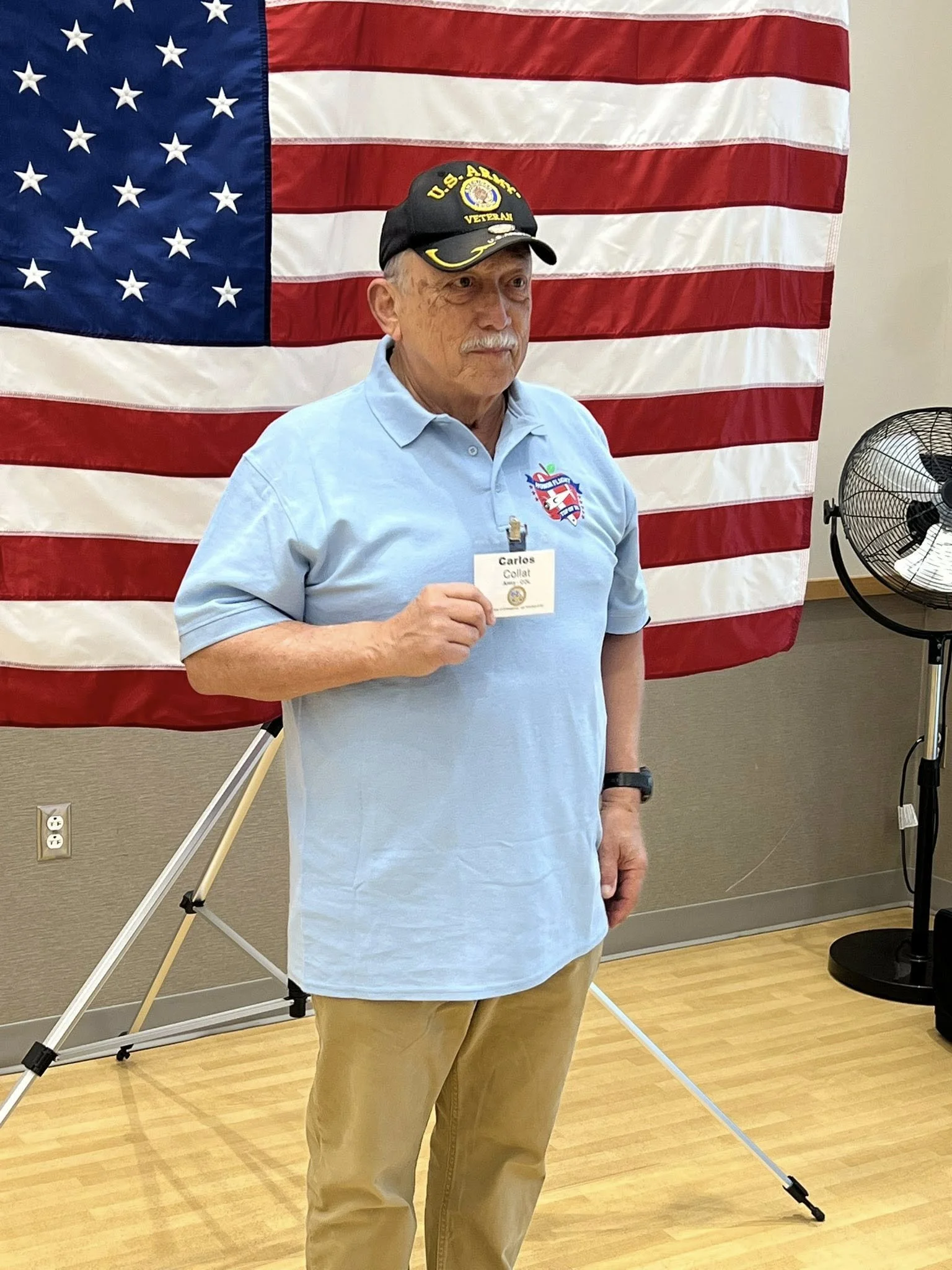 An elderly man standing indoors in front of a large American flag. He is wearing a black cap with military and veteran insignia, a light blue polo shirt with a logo and name tag, and tan pants. He is holding a small object in his right hand and has a