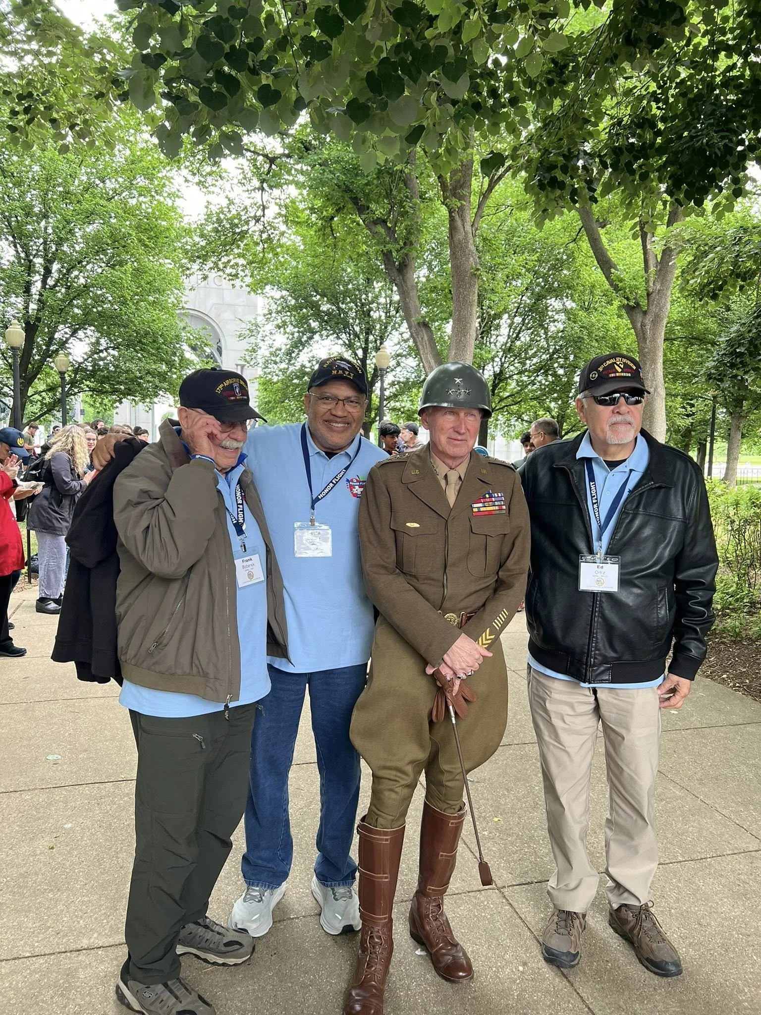 Four men standing together outdoors, surrounded by green trees and other people. The second man from the right is dressed in a military uniform with medals. The men are smiling or looking serious, and some are wearing caps or sunglasses.