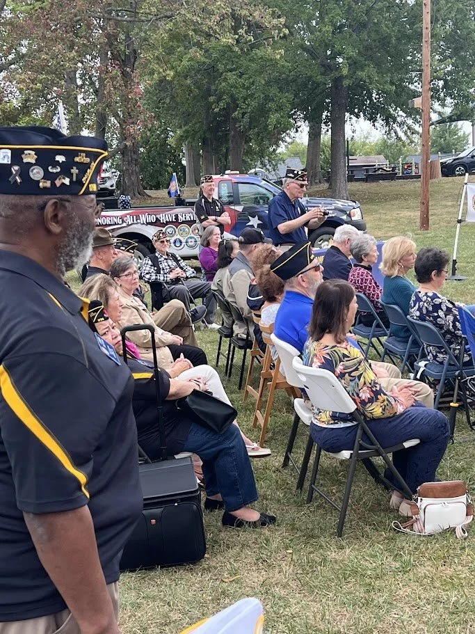 A gathering of veterans, some wearing military hats, seated outdoors on folding chairs at a memorial service or patriotic event. A man in uniform is in the foreground, with a police vehicle and a memorial display in the background.