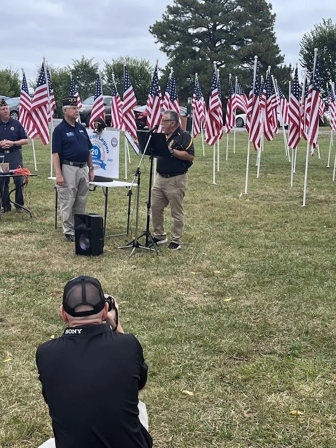 A group of men standing outdoors with American flags behind them. One man is speaking at a microphone, and another man is reading from a paper. A person in the foreground is taking a photo or recording with a Sony camera.
