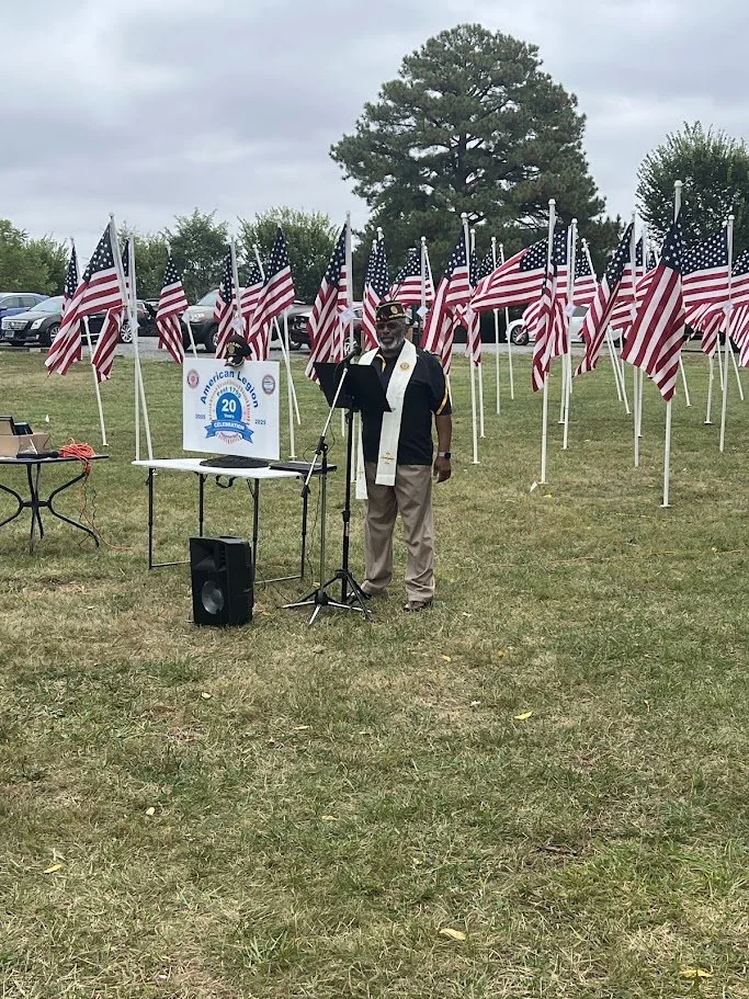 A man standing at a microphone outdoors, surrounded by American flags, with a sign commemorating the 20th anniversary of a patriotic organization, and parked cars in the background.