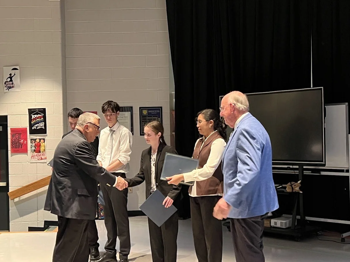 A group of five people, including two young men and three women, standing in a line in an indoor setting, with one person shaking hands with the individual in front of him, during a formal event or award ceremony.