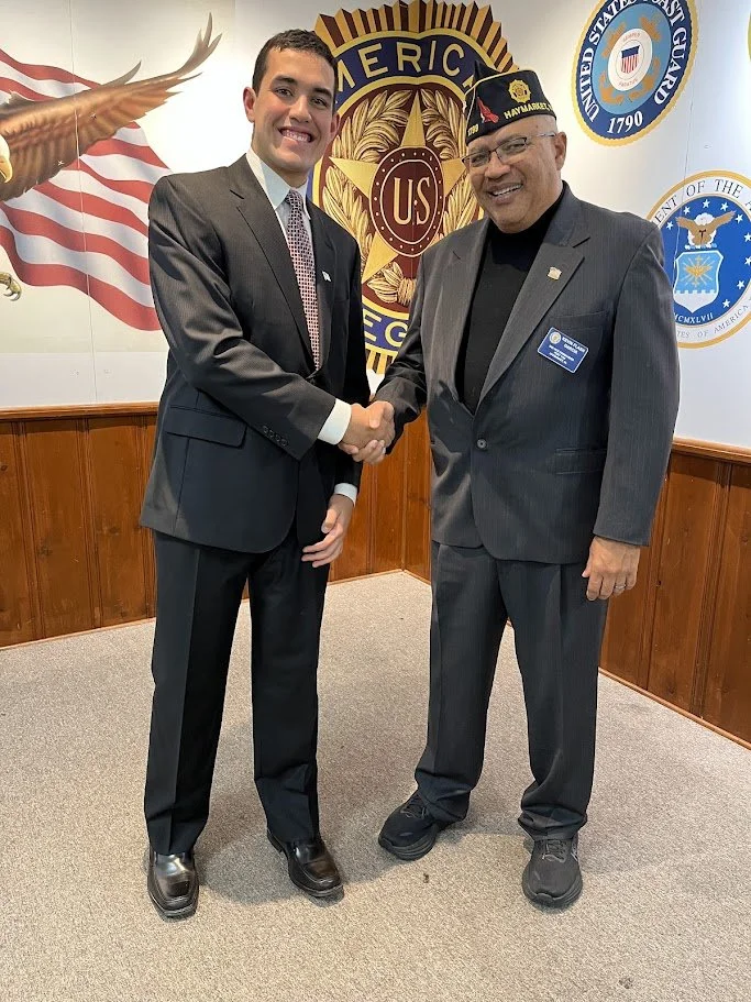 Two men in suits shaking hands inside a room with veteran and patriotic banners and emblems on the wall.