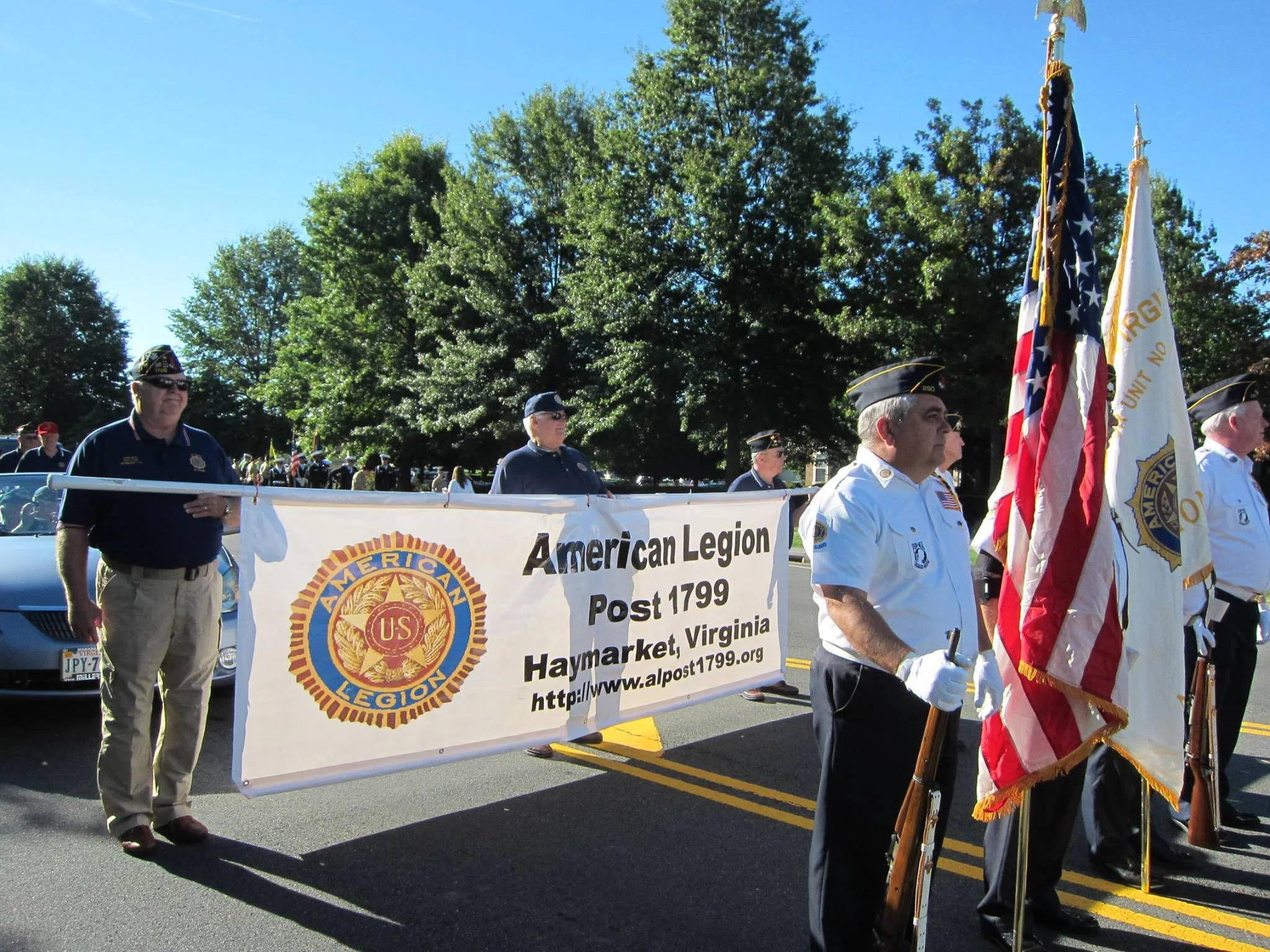 Veterans and members of the American Legion standing at attention during a parade, holding American flags and a banner for Post 1799 in Haymarket, Virginia, with trees and spectators in the background.