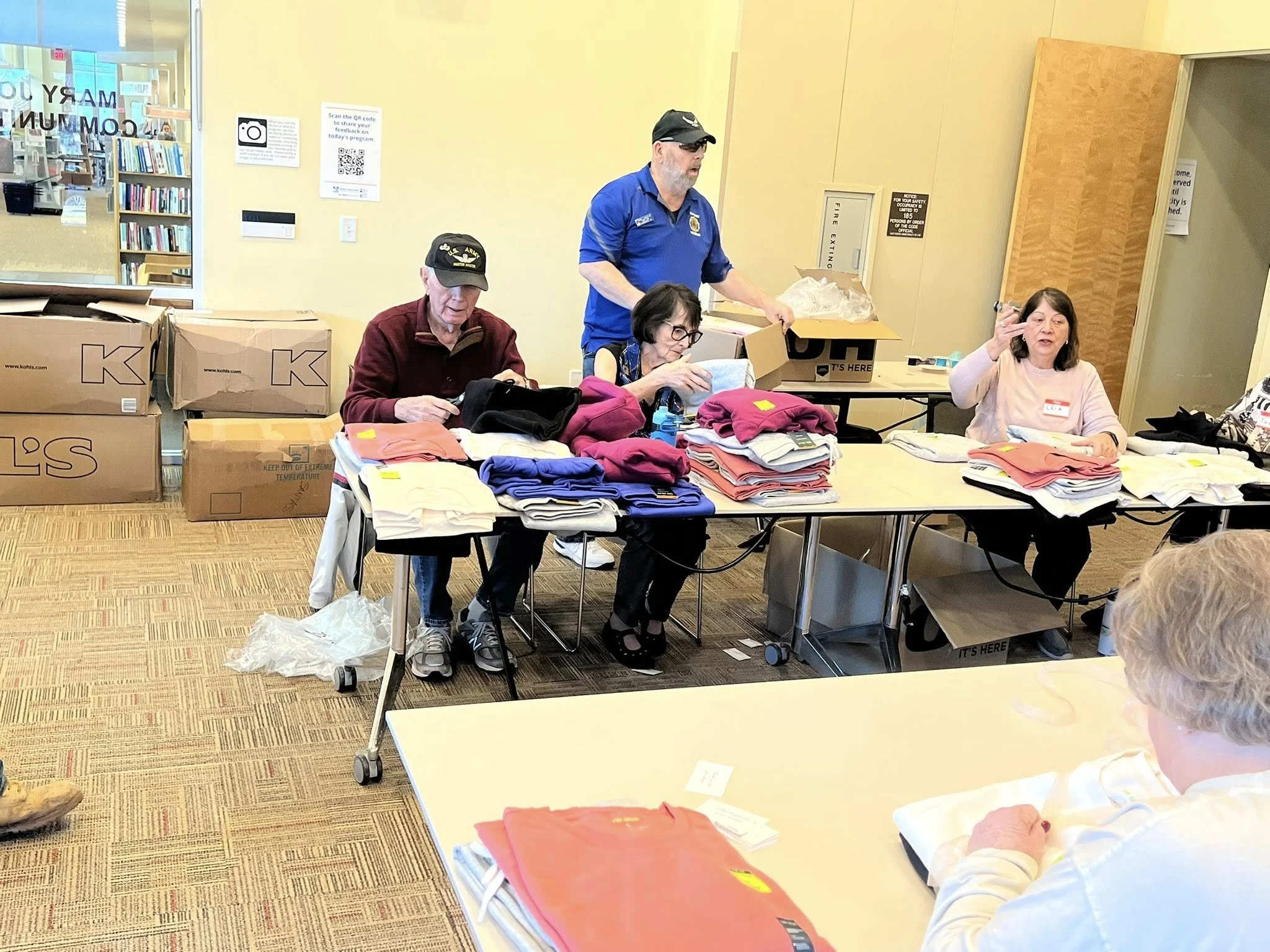 A group of five people working at a table in a library, sorting and organizing folded clothing and sweatshirts.