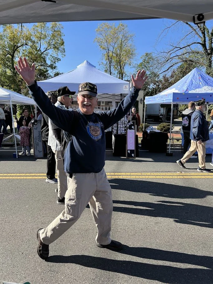 A smiling man with glasses and a cap, wearing a navy blue sweatshirt and beige pants, standing on a street with his arms raised and spread wide, at an outdoor event with tents and trees in the background.
